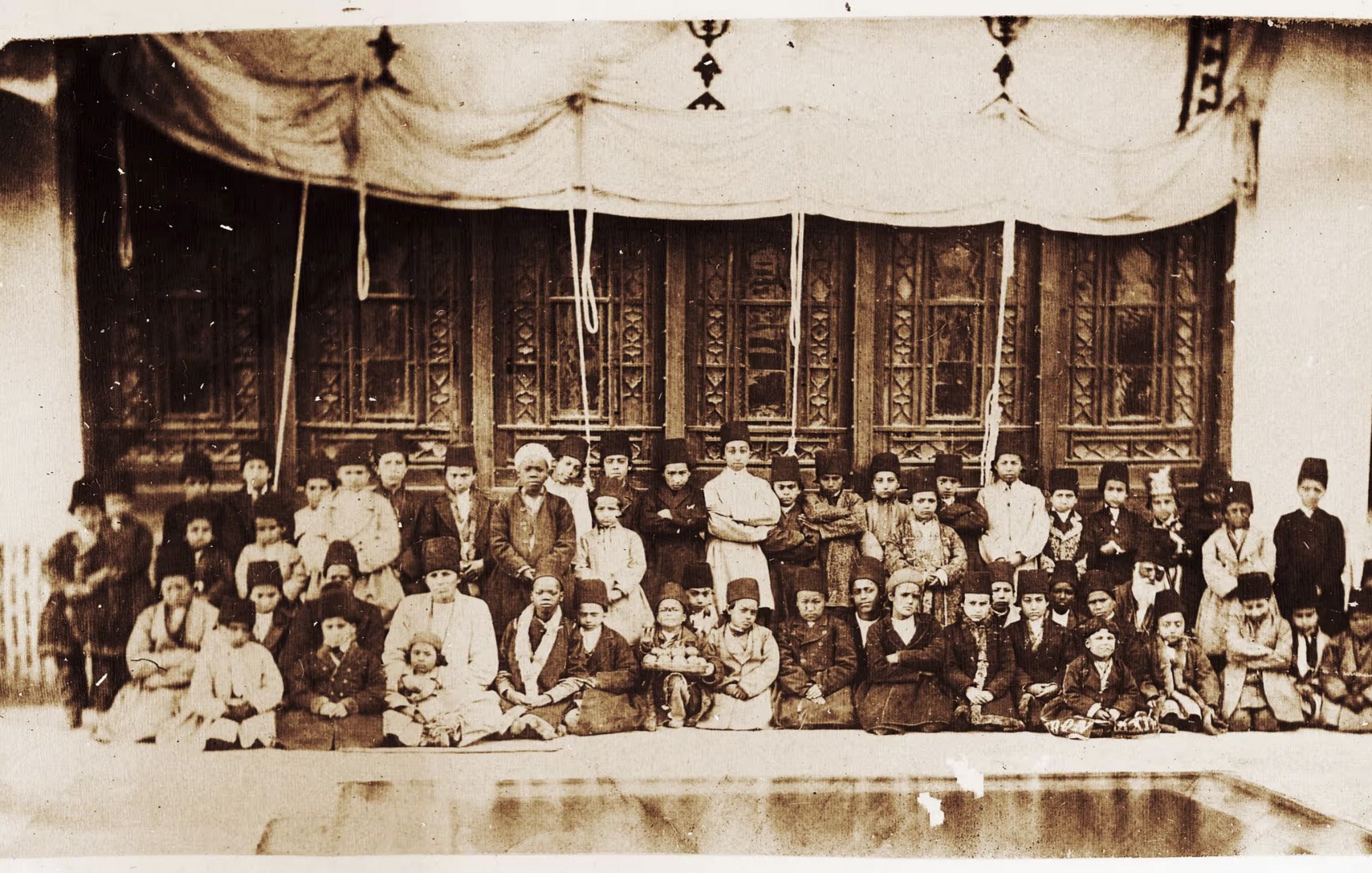 L'image présente un groupe de personnes, probablement une photographie en noir et blanc, où l'on peut voir une grande assemblée d'hommes, de femmes et d'enfants. Ils sont assis ou debout devant un bâtiment avec des fenêtres ornées et des décorations. Les vêtements portés semblent typiques d'une époque passée, suggérant un contexte historique. L'ensemble donne une impression de communauté ou d'événement social.