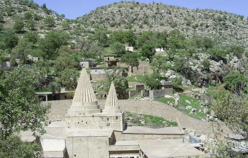 L'image montre un paysage montagneux avec une structure architecturale distinctive au centre. Cette structure présente deux tours ou pinacles coniques, typiques d'un style architectural particulier. Autour, on aperçoit des bâtiments en pierre et en béton, ainsi que des arbres verdoyants qui ajoutent une touche de nature. Le terrain est légèrement accidenté, avec des collines à l'arrière-plan, créant un environnement pittoresque et tranquille.