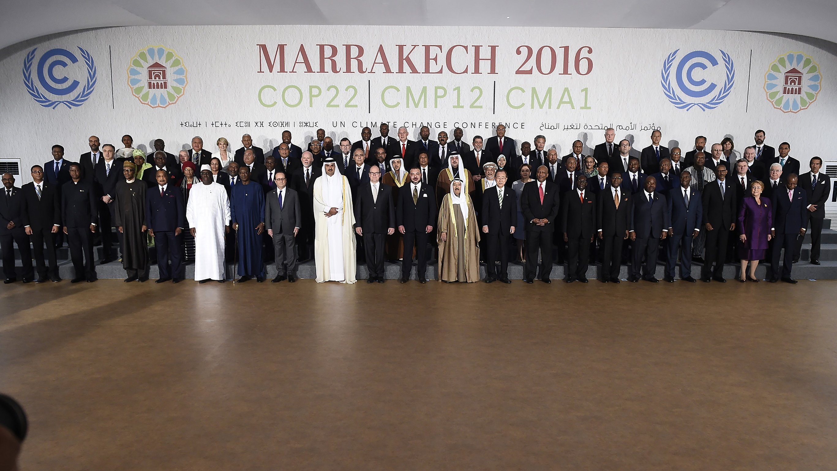 L'image montre un groupe de participants rassemblés lors de la conférence COP22 qui s'est tenue à Marrakech en 2016. Les membres représentés incluent des dirigeants et des délégués de divers pays, vêtus de costumes formels et de tenues traditionnelles. Ils se tiennent debout sur des gradins, souriant pour la photo officielle, avec un fond affichant le nom de l'événement et des logos des Nations Unies. L'ambiance évoque un sentiment de collaboration internationale pour faire face aux défis climatiques.