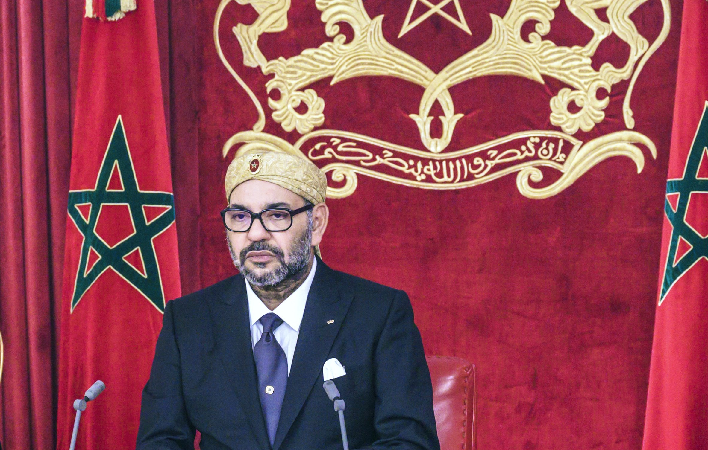 L'image montre un homme assis à un bureau, habillé élégamment en costume noir avec une cravate. Il porte un turban ou un couvre-chef traditionnel. En arrière-plan, on peut voir deux drapeaux marocains ornés d'une étoile à cinq branches. Le décor est riche, avec un fond rouge et une représentation du blason du Maroc. L'homme semble concentré et prêt à s'exprimer.