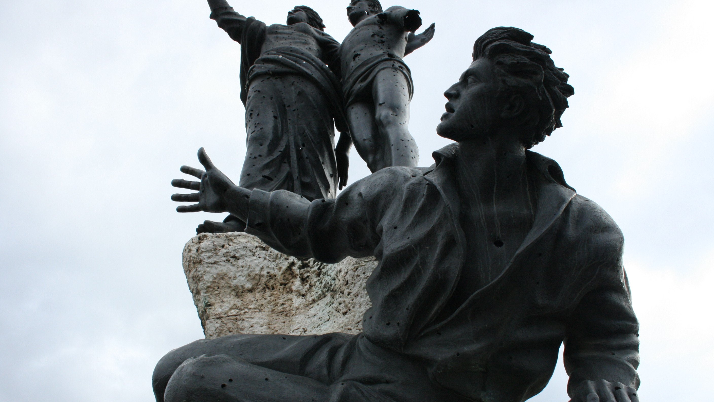 L'image montre une sculpture en bronze représentant plusieurs figures humaines. Au premier plan, un homme est assis, les bras ouverts, avec une expression qui semble transmettre une émotion forte. Derrière lui, deux figures debout sont également visibles, levant les bras, ce qui pourrait suggérer un message de célébration ou de libération. La sculpture est placée sur une base rocheuse, ajoutant une dimension texturisée à l'ensemble. Le ciel apparaît nuageux, ce qui donne un ton dramatique à la scène.