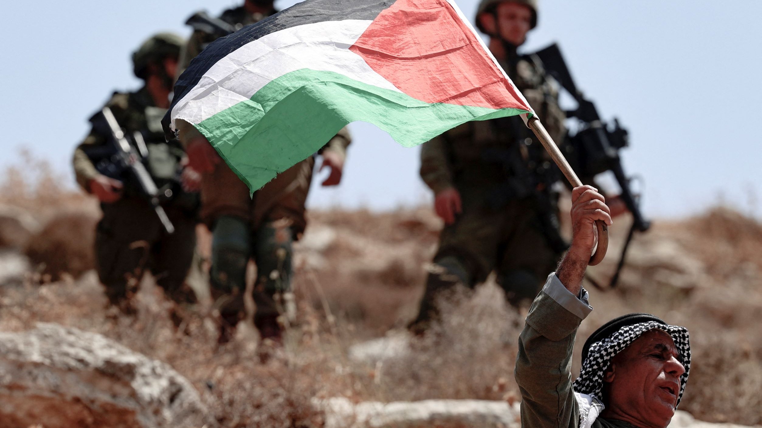 L'image montre un homme brandissant fièrement un drapeau palestinien. Il semble être en première ligne, tandis que des soldats en arrière-plan, probablement des soldats israéliens, sont visibles. Le paysage est sec et rocailleux, suggérant un environnement rural ou semi-aride. L'atmosphère semble tendue en raison de la présence militaire et du geste de protestation de l'homme.