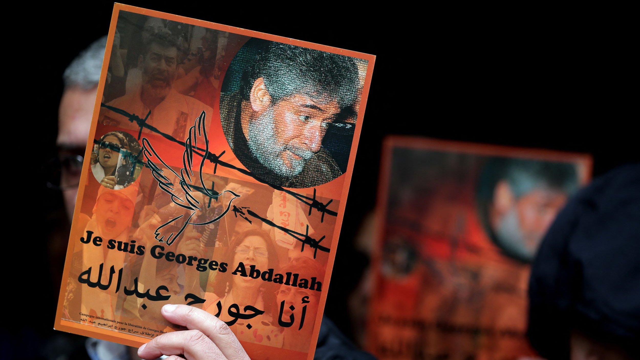 L'image montre une personne tenant une affiche. L'affiche met en avant le message "Je suis Georges Abdallah" en français et en arabe. Elle inclut un portrait de Georges Abdallah entouré de motifs symboliques, comme des barbelés et une colombe. En arrière-plan, on aperçoit des visages et des motifs qui suggèrent un contexte de soutien et d'engagement. L'ambiance semble être celle d'une manifestation ou d'un rassemblement pour la défense de ses droits.