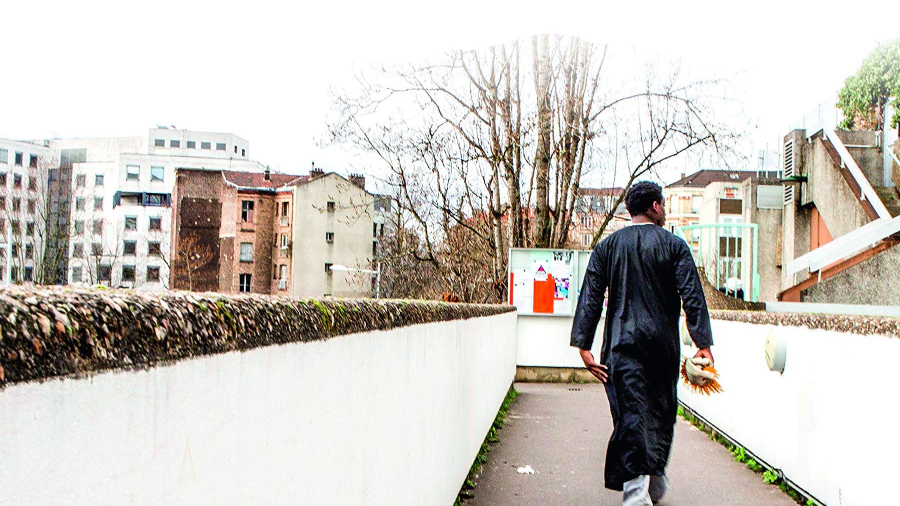 L'image montre un homme marchant sur un chemin urbain. Il porte une tenue traditionnelle, une sorte de longue robe noire. En arrière-plan, on peut voir des bâtiments modernes et anciens, ainsi que des arbres dépouillés. L'environnement semble calme, avec une lumière diffuse, peut-être un jour nuageux. L'homme avance vers l'horizon, de dos, ce qui donne une impression de solitude et de contemplation.