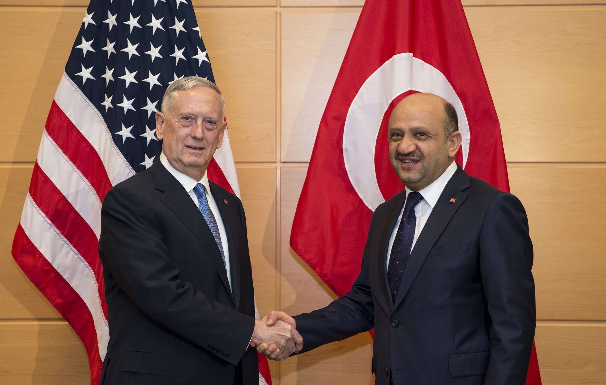 L'image montre deux hommes se serrant la main, probablement en signe de coopération ou d'accord. À l'arrière-plan, on peut voir deux drapeaux : celui des États-Unis à gauche et celui de la Turquie à droite. Les deux hommes portent des costumes formels, et l'ambiance semble être celle d'une rencontre officielle.