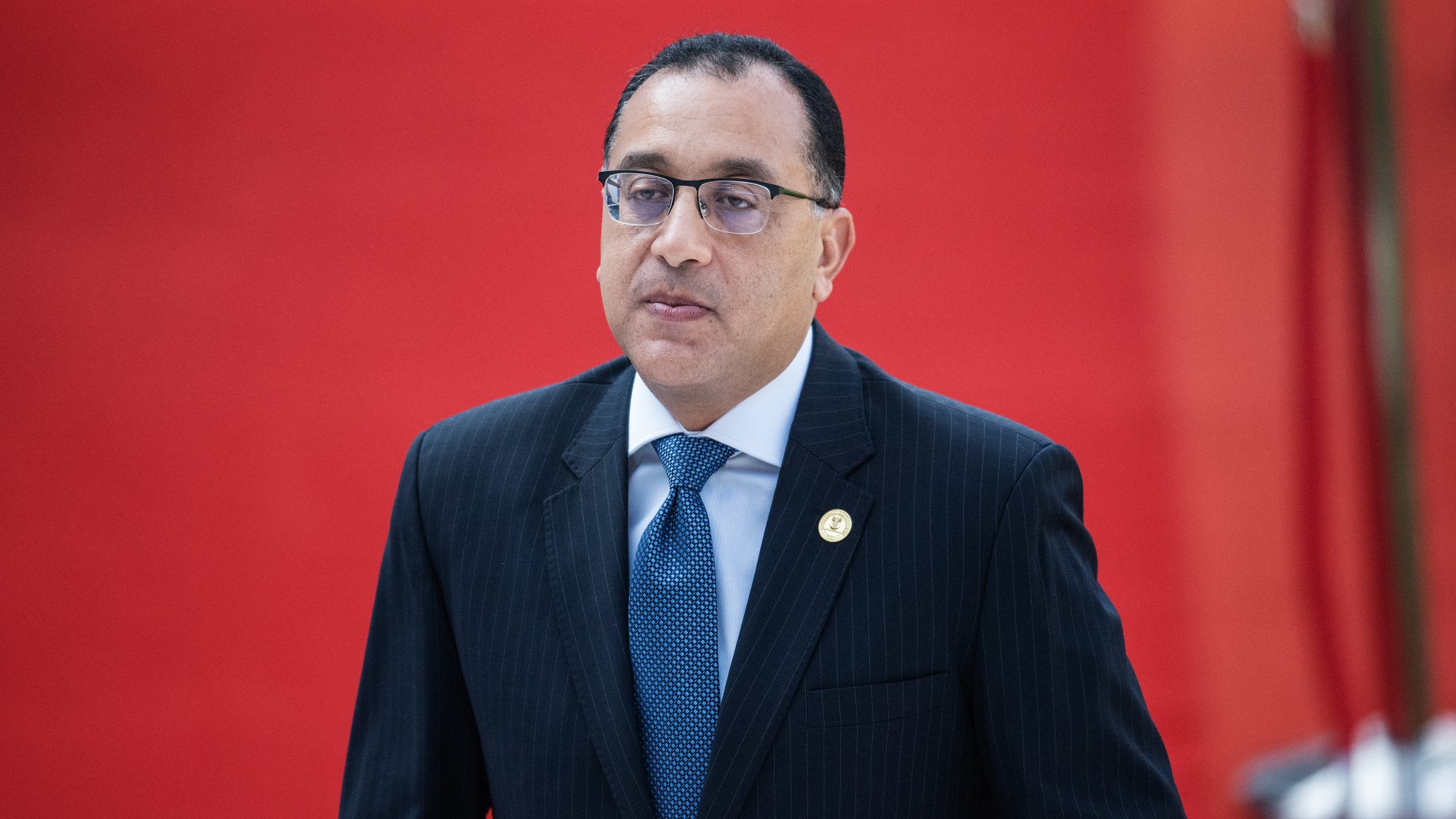 L'image montre un homme en costume noir avec une chemise blanche et une cravate bleue. Il porte des lunettes et a une expression sérieuse. L'arrière-plan est rouge, ce qui crée un contraste vif avec sa tenue. L'homme semble être dans un cadre officiel ou lors d'un événement important.