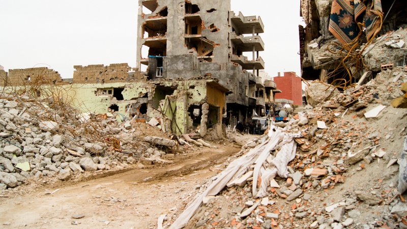 L'image montre un paysage urbain dévasté par la guerre ou une catastrophe. On peut voir des bâtiments partiellement détruits, présentant des murs troués et des structures effondrées. Des débris encombrent le sol, et des morceaux de béton et des matériaux de construction sont éparpillés partout. La rue semble désolée, soulignant l'impact de la destruction sur l'environnement urbain. Les couleurs sont sombres et tristes, renforçant le sentiment de désespoir et de désolation.