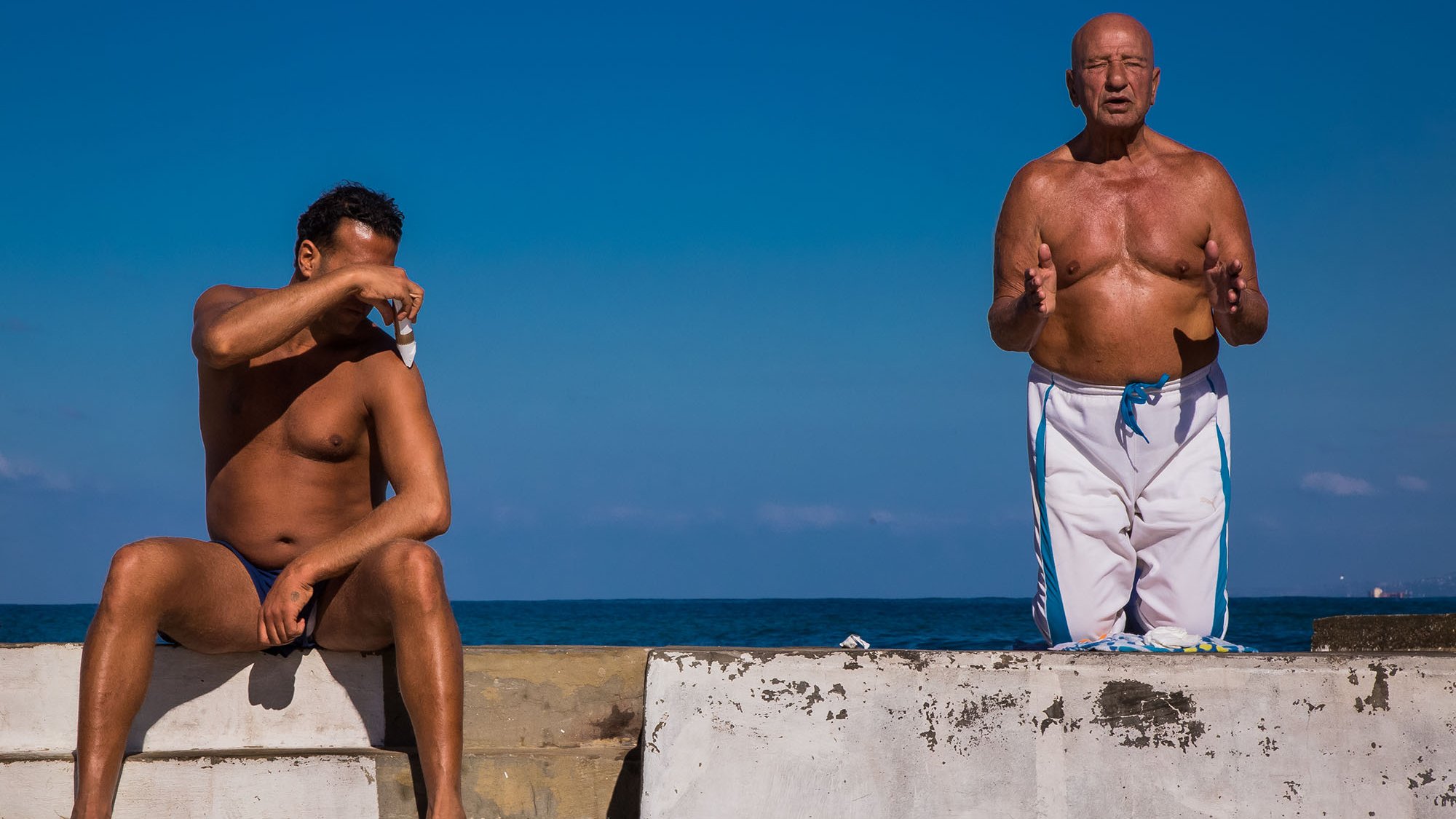 L'image montre deux hommes sur une structure en béton au bord de l'eau. L'un des hommes est assis, torse nu, avec les bras croisés sur un genou, semblant protéger ses yeux du soleil. L'autre homme, debout à côté de lui, est également torse nu et semble parler ou gesticuler, comme s'il était en pleine conversation. En arrière-plan, on peut voir un ciel bleu clair et une étendue de mer, créant une atmosphère estivale et décontractée.