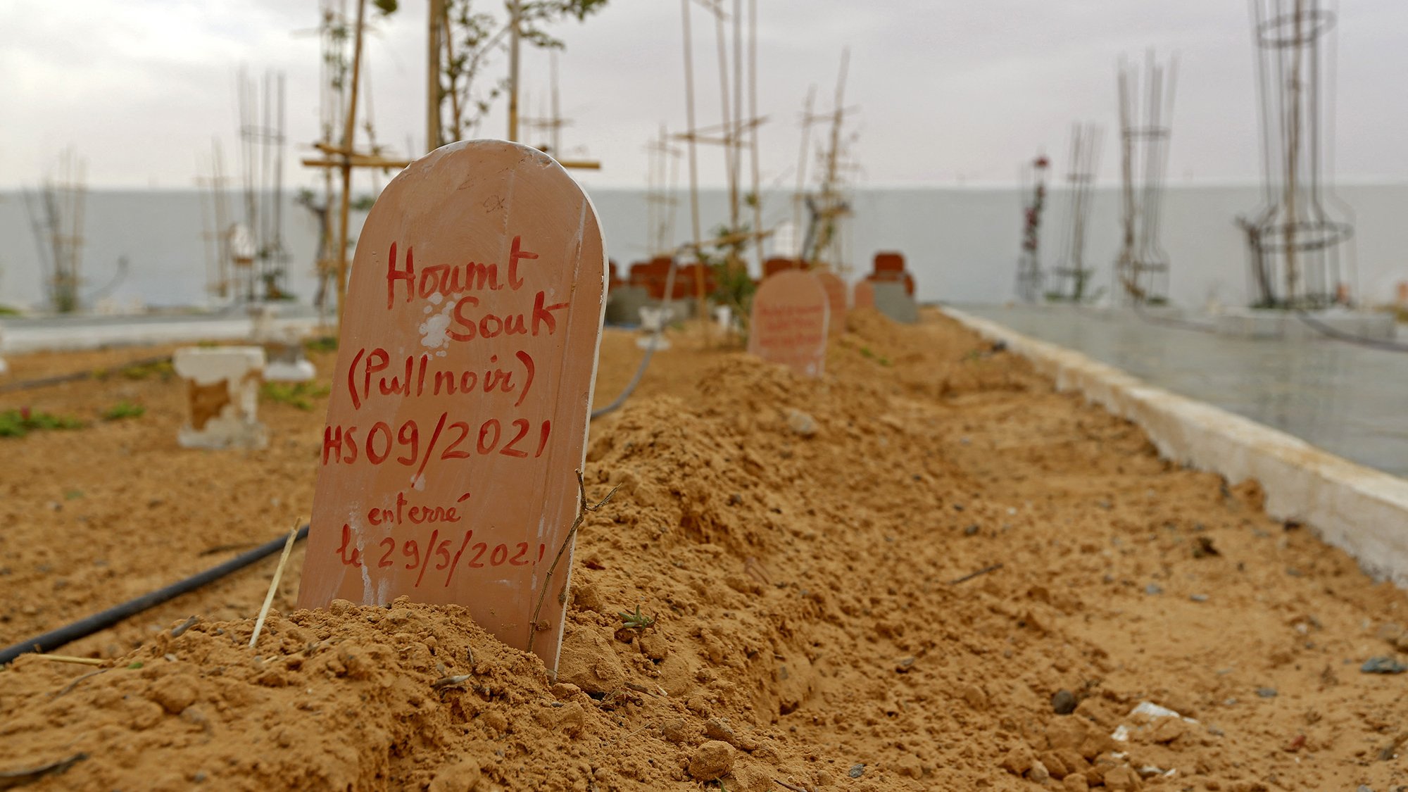 L'image montre un cimetière avec des tombes marquées par des plaques en terre cuite. Le sol est sablonneux et légèrement irrégulier, avec quelques plantes éparses. En arrière-plan, on remarque des structures en construction, probablement des bâtiments ou des monuments. Le ciel semble nuageux, créant une atmosphère calme mais mélancolique. Les inscriptions sur les plaques indiquent les noms et des dates, suggérant que ces êtres chers ont été récemment enterrés.