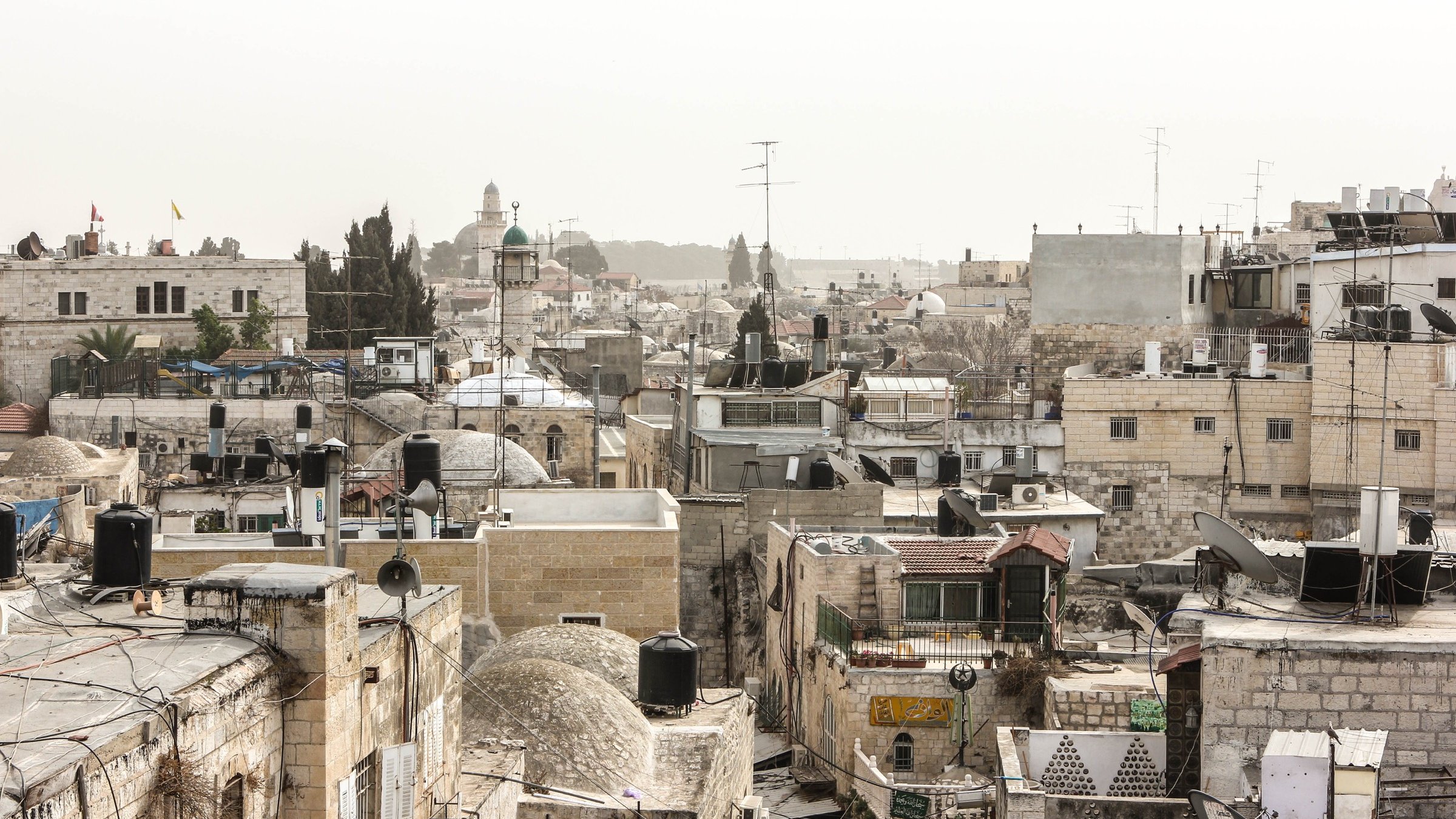 L'image montre un paysage urbain d'une ville ancienne, probablement Jérusalem. On peut voir une multitude de bâtiments en pierre avec des toits en tôle et des dômes, créant une texture variée. Les structures sont serrées les unes contre les autres, et quelques antennes de télévision sont visibles. Au loin, se distingue un minaret ou une coupole, indiquant la présence d'une mosquée. L'atmosphère semble un peu brumeuse, donnant une impression de chaleur et d'ancienneté à la scène.