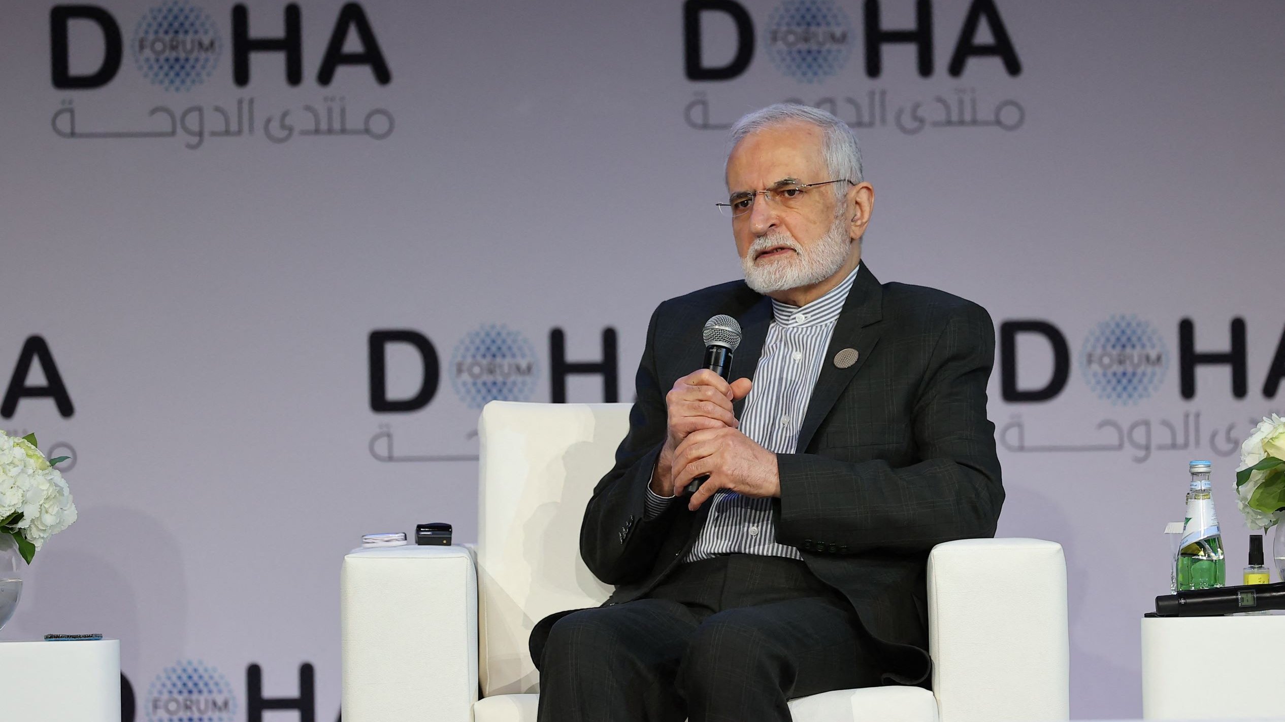 L'image montre un homme assis sur un fauteuil blanc, participant à une conférence ou un forum. Il porte une tenue formelle et semble être en train de s'exprimer ou d'écouter. En arrière-plan, on peut voir un logo avec le mot "DOHA" et d'autres inscriptions en arabe. Des fleurs et une bouteille d'eau sont également visibles sur la scène. L'atmosphère semble professionnelle et tranquille.