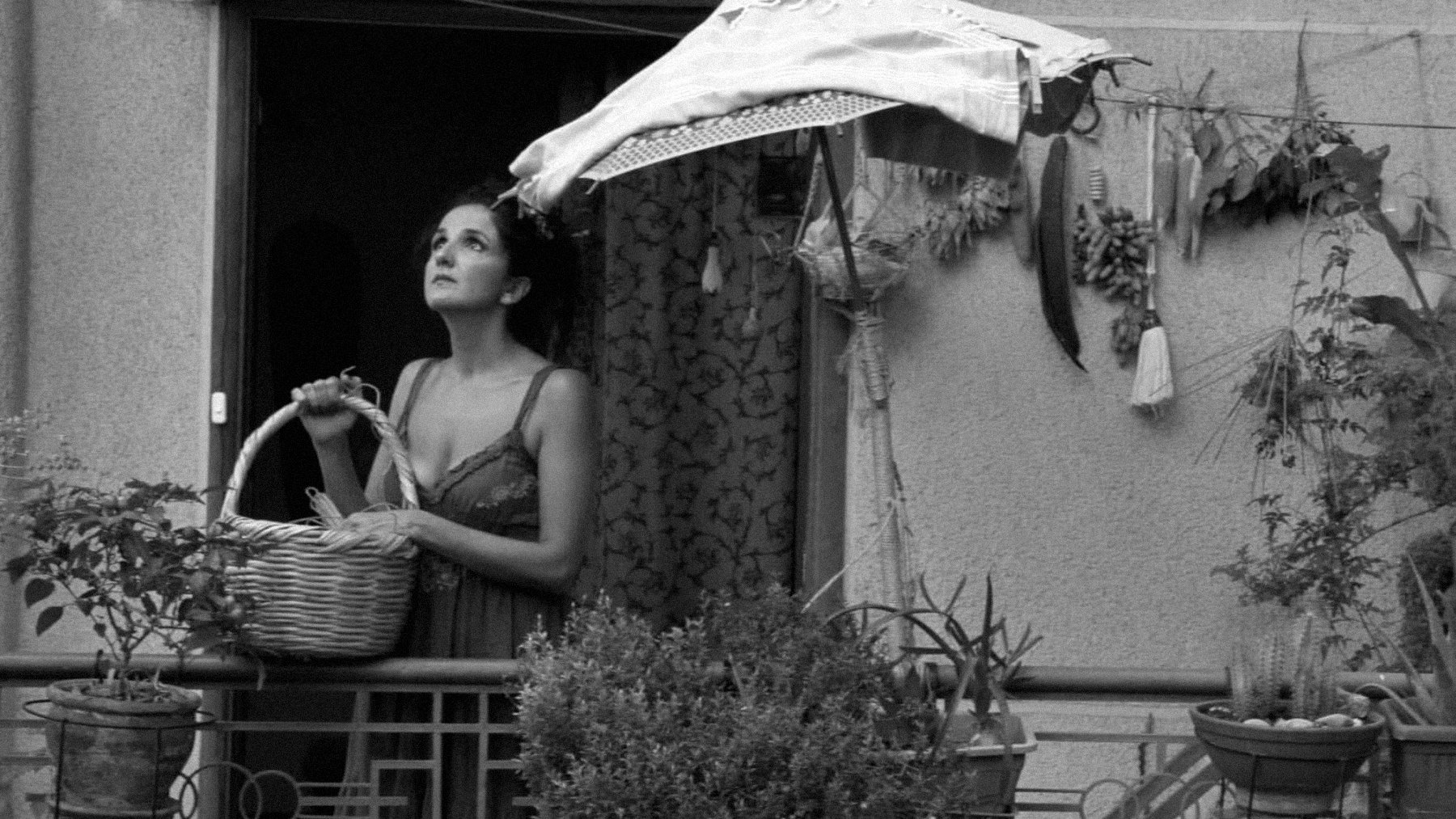 L'image montre une femme se tenant sur un balcon, tenant un panier dans ses bras. Elle semble contemplative, regardant au loin. Le balcon est décoré de plusieurs plantes et herbes suspendues. Un parasol en toile protège en partie la femme du soleil. L'image est en noir et blanc, ce qui lui donne une atmosphère nostalgique et mélancolique.