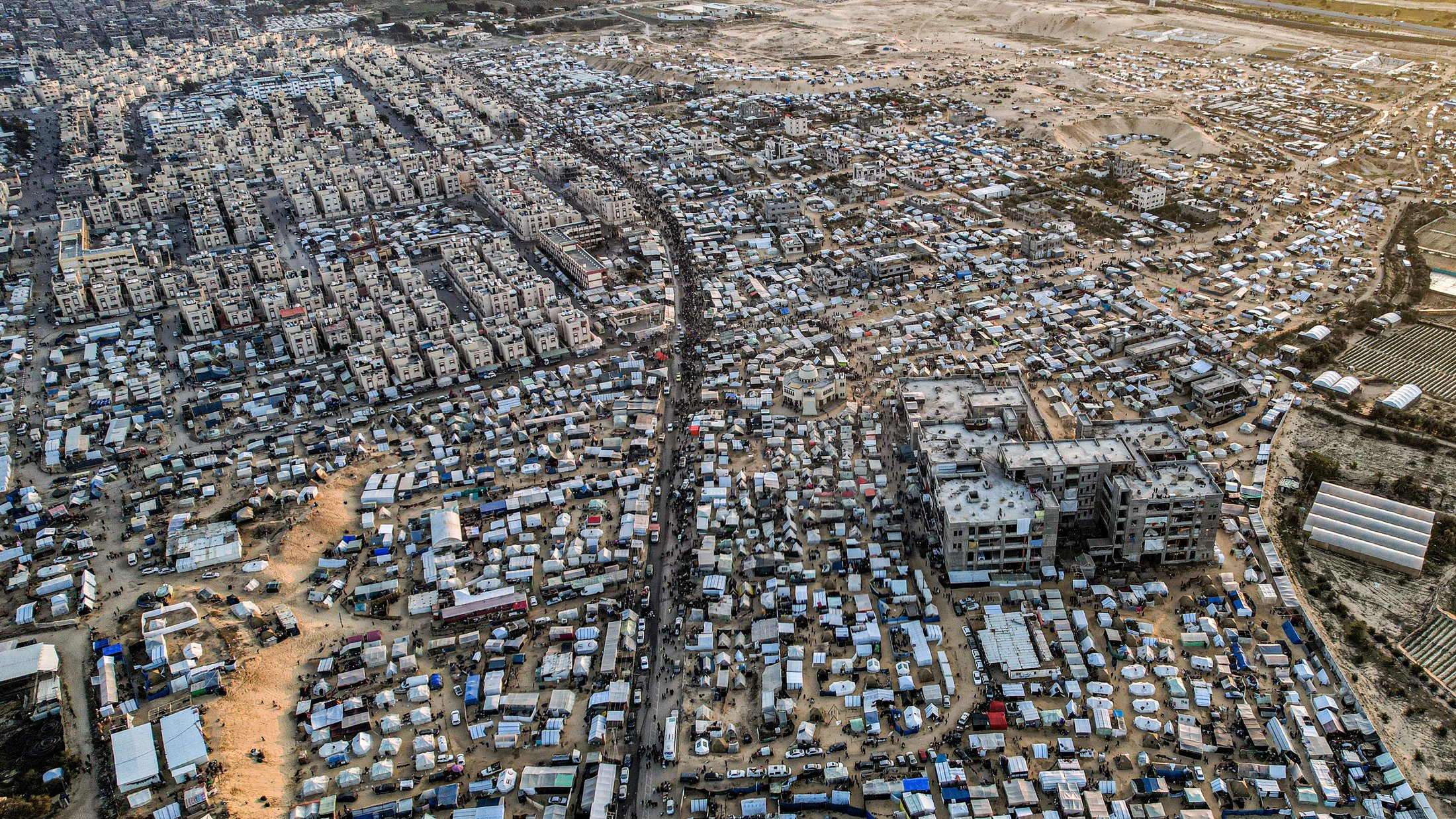 L'image montre une vue aérienne d'une vaste zone urbaine très densément peuplée, probablement une ville en développement ou un bidonville. On peut distinguer des habitations de différentes tailles et styles, de nombreuses structures temporaires, ainsi que des routes et des chemins qui traversent l'ensemble de la zone. La scène est marquée par un mélange de constructions durables et de tentes, suggérant une forte densité de population ainsi que des conditions de vie variées. En arrière-plan, on aperçoit des terrains plus dégagés, peut-être des zones agricoles ou industrielles.