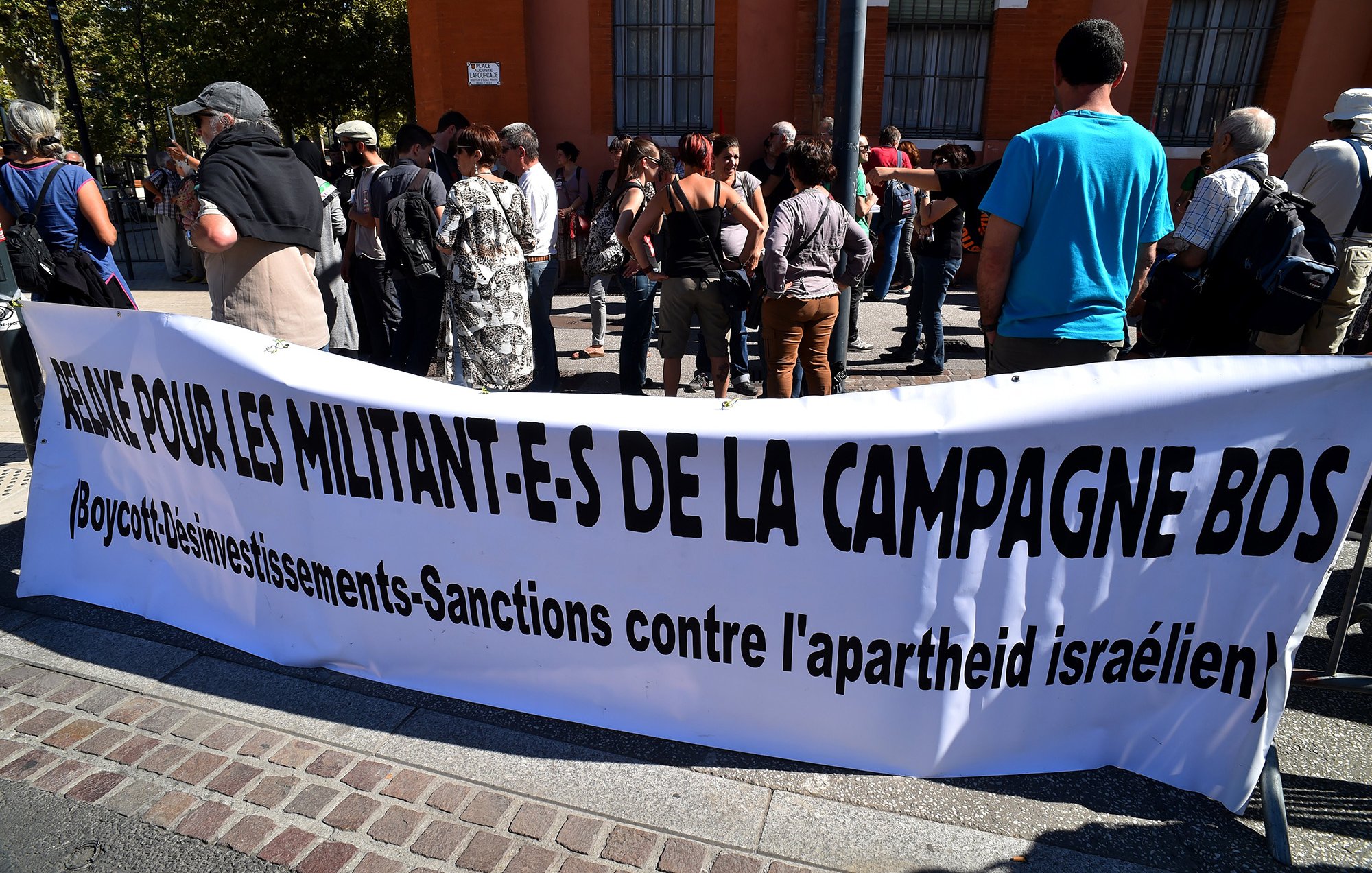 L'image montre un groupe de personnes rassemblées devant une bannière. La bannière, qui est en français, demande un soutien pour les militants de la campagne BDS, qui signifie Boycott, Désinvestissement et Sanctions contre l'apartheid israélien. Les manifestants semblent engagés et attentifs, participant à une action de protestation. Le décor urbain en arrière-plan indique que l'événement se déroule dans une ville, probablement en plein air.