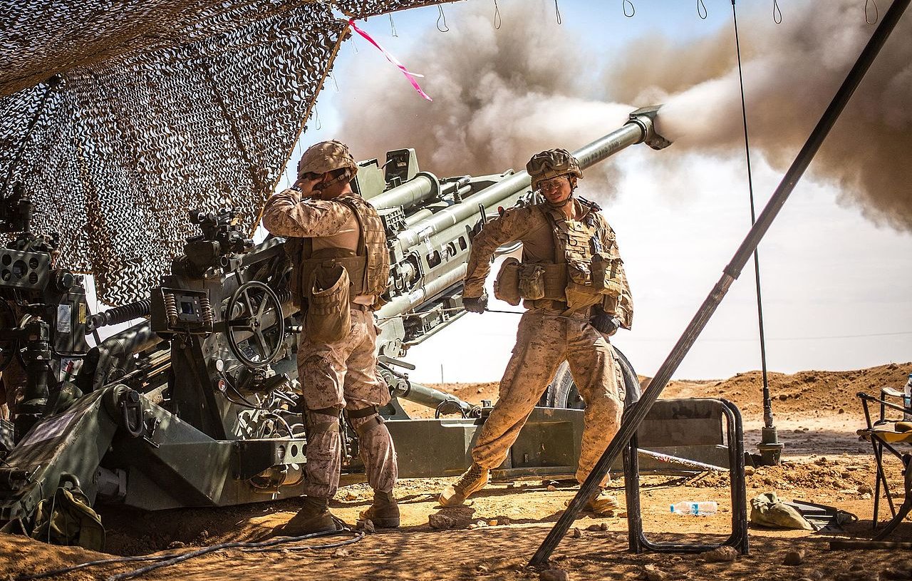 L'image montre deux soldats en uniforme militaire, probablement dans un environnement désertique. Ils sont proches d'une grande pièce d'artillerie qui est en train de tirer, créant un nuage de fumée. L'un des soldats semble gérer l'équipement tandis que l'autre se tient debout, surveillant la situation. Un camouflage est utilisé pour dissimuler l'équipement militaire, et le paysage environnant est aride avec un ciel dégagé. L'atmosphère semble tendue et concentrée, rappelant des opérations militaires.