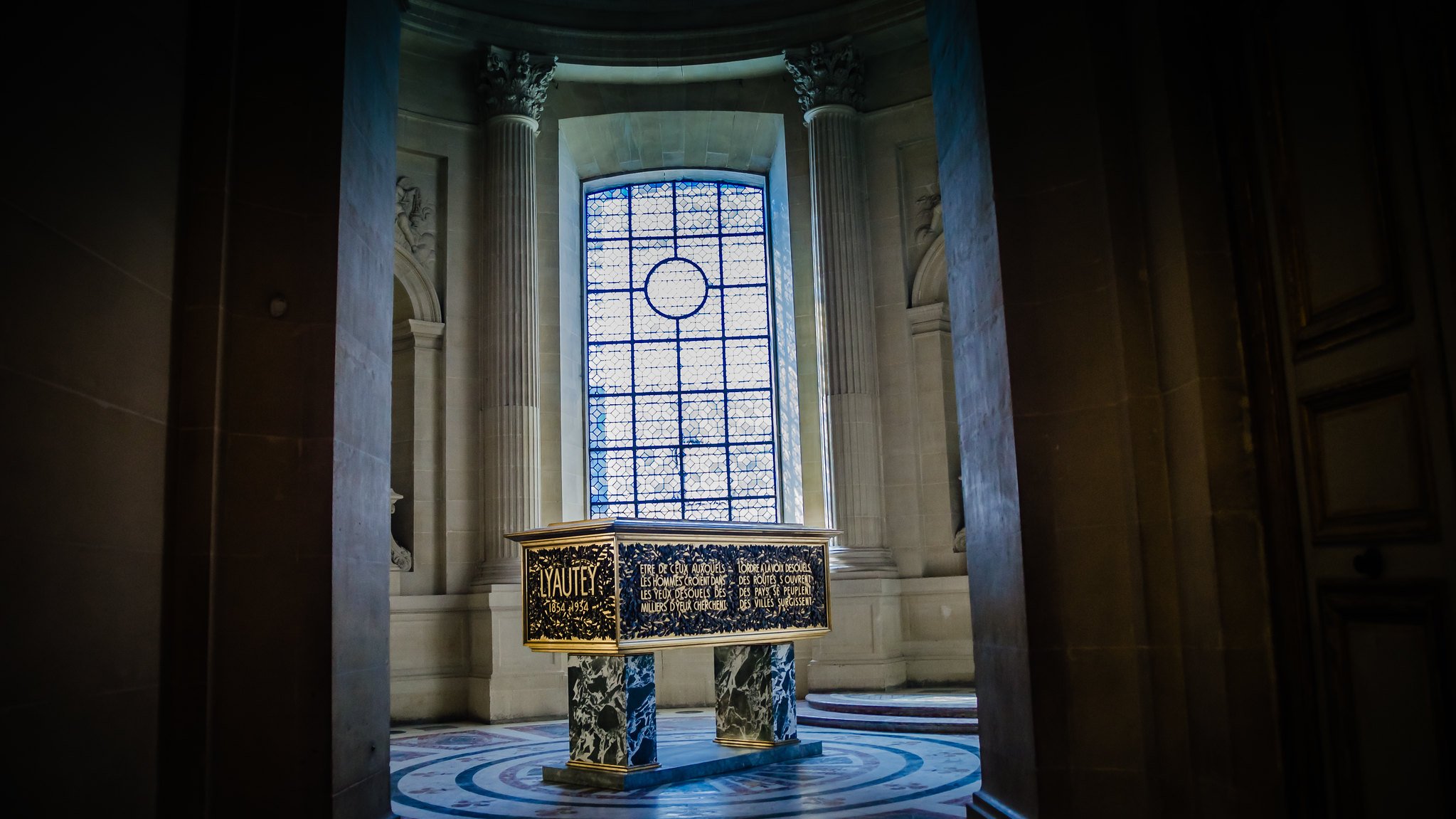 L'image montre une pièce intérieure d'un bâtiment majestueux, probablement un palais ou un musée. Au centre, il y a un grand meuble ou une caisse ornée de motifs décoratifs dorés et peints. Les murs sont en pierre, avec de très grandes fenêtres, à travers lesquelles entre une lumière douce, créant une atmosphère solennelle. Les colonnes et le sol richement décoré ajoutent à la grandeur de l'espace.