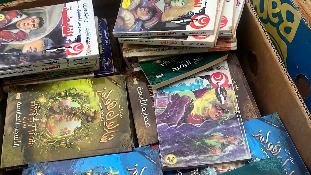 L'image montre une boîte remplie de livres empilés les uns sur les autres. On peut apercevoir différents titres en arabe sur les couvertures, illustrés par des images colorées. Certains livres semblent appartenir à des séries de contes ou de romans graphiques, avec des illustrations captivantes et des textes en arabe. L'ensemble donne une impression d'abondance littéraire.