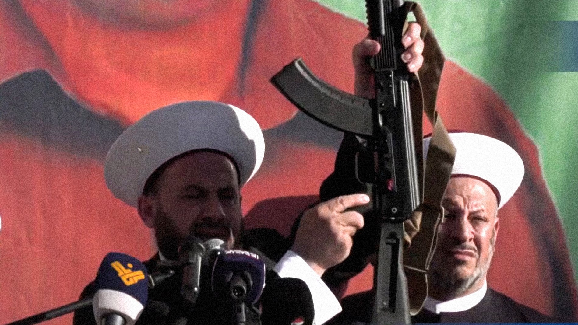 L'image montre deux hommes portant des chapeaux traditionnels, se tenant devant un grand panneau ou une affiche représentant une figure politique ou historique. L'un des hommes brandit une arme, probablement un fusil d'assaut, en faisant un geste qui attire l'attention du public. L'atmosphère semble solennelle et les deux hommes semblent s'adresser à une foule, ce qui suggère un contexte d'engagement politique ou religieux.