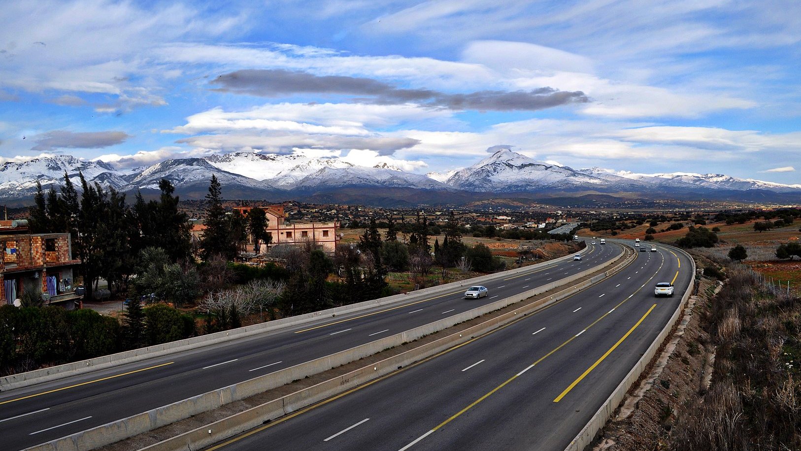 L'image montre une route large et bien entretenue, qui s'étend à travers un paysage pittoresque. On peut voir des montagnes majestueuses au loin, avec des sommets enneigés qui contrastent avec le ciel bleu parsemé de nuages. De chaque côté de la route, il y a quelques bâtiments et de la végétation, ce qui ajoute à la beauté de la scène. Cette vue évoque une ambiance paisible et naturelle, avec un mélange de modernité et de paysages ruraux.