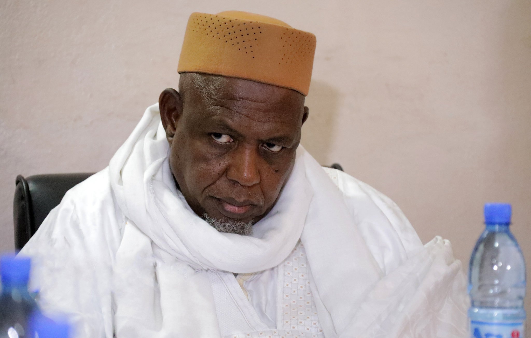 L'image montre un homme, probablement assis, portant une tenue traditionnelle, incluant un chapeau de couleur beige et un long habit blanc. Son expression semble sérieuse et réfléchie, tandis qu'il regarde dans une direction avec une certaine intensité. En arrière-plan, on peut voir des bouteilles d'eau sur une table. L'environnement semble simple, suggérant un cadre formel ou communautaire.