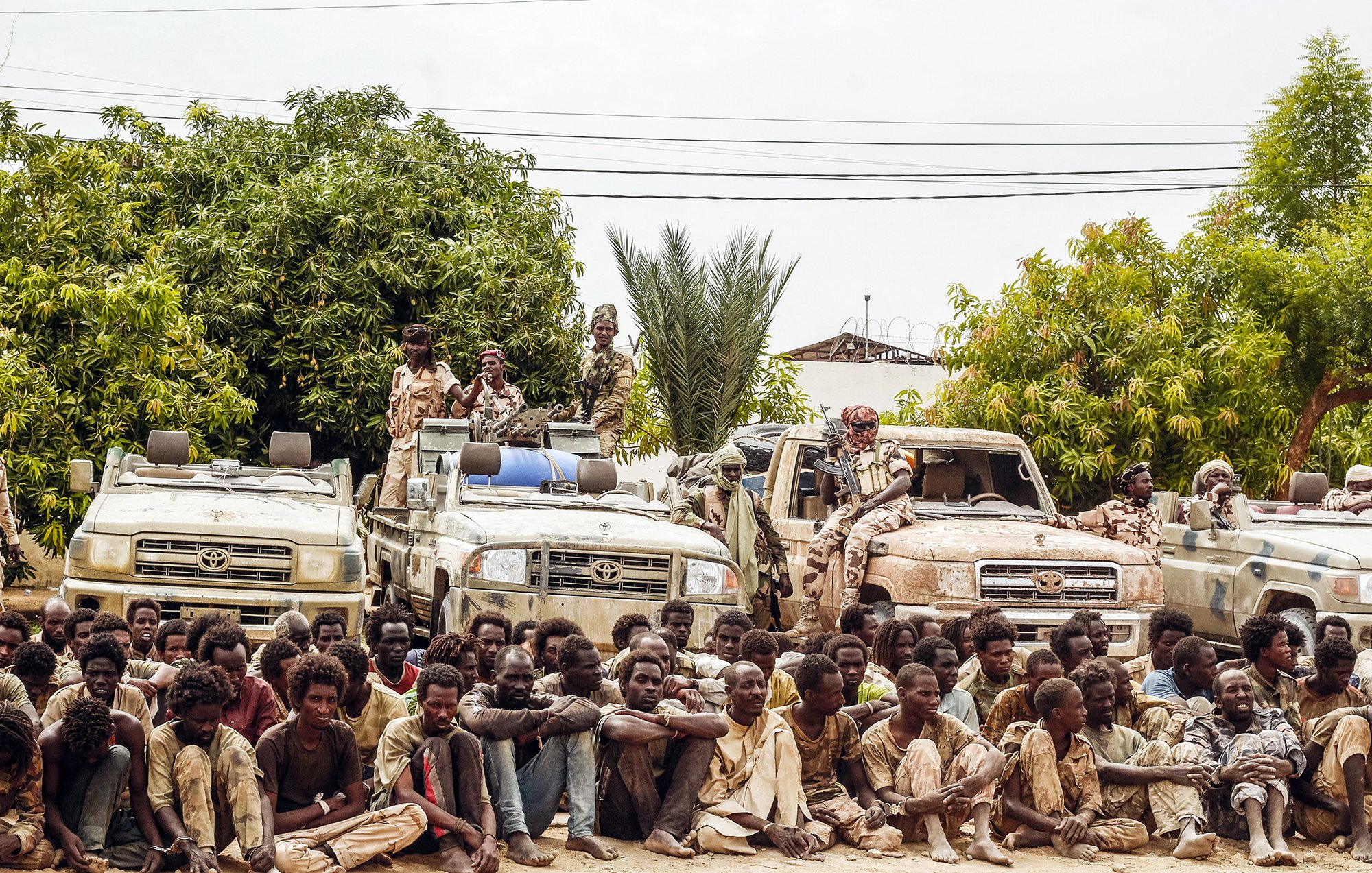 L'image montre un groupe de personnes assises au sol, semblant fatiguées et dans des vêtements usés, tandis qu'elles sont entourées de plusieurs véhicules tout-terrain. À l'arrière-plan, on peut voir des arbres et des plantes, suggérant un environnement rural. Certains individus semblent garder les véhicules, et l'atmosphère générale donne une impression de tension ou de détresse.