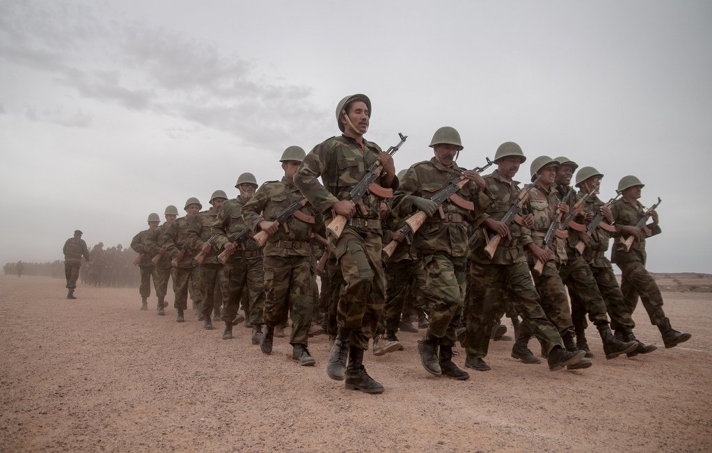 L'image représente un groupe de soldats marchant en formation sur un chemin poussiéreux. Ils sont habillés en tenue militaire camouflée et portent des casques. Chaque soldat tient un fusil à la main. L'atmosphère semble sérieuse, et le ciel est nuageux, suggérant peut-être des conditions climatiques difficiles. En arrière-plan, on aperçoit une ligne de végétation ou d'arbres, ajoutant à l'impression d'un environnement militaire.