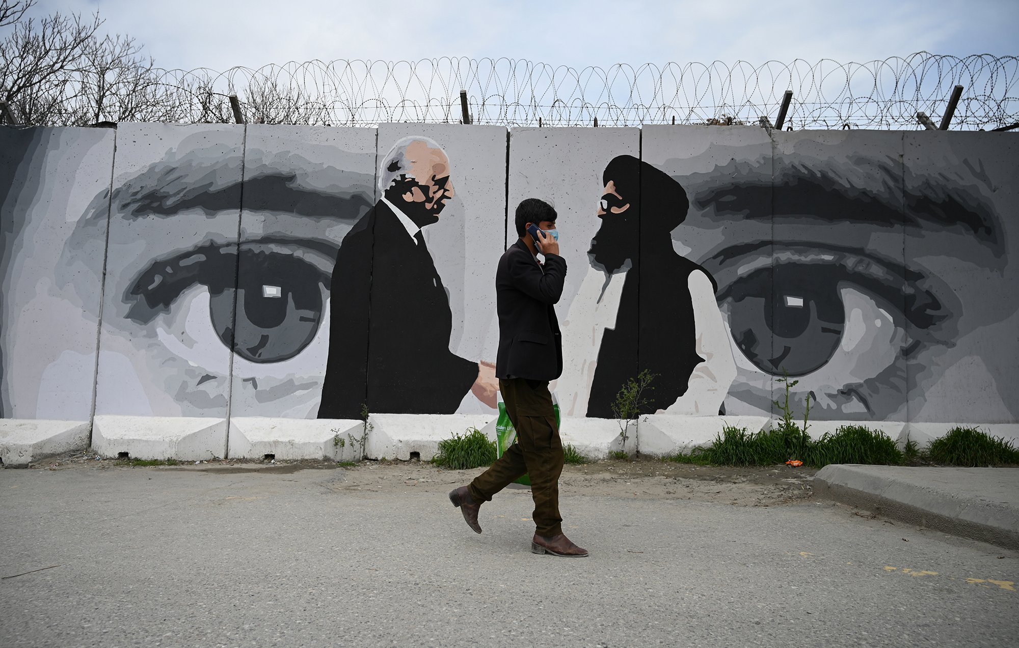 L'image montre un homme marchant sur une route, portant un costume sombre, tandis qu'il téléphone. En arrière-plan, on aperçoit un mur décoré d'une fresque qui représente des visages stylisés, probablement en noir et blanc. L'œuvre met en avant des yeux perçants et deux silhouettes, ce qui semble évoquer des figures significatives. La scène dégage une atmosphère de tension et de réflexion, juxtaposition entre le monde urbain et l'art mural. Des barbelés sur le mur ajoutent une dimension de restriction ou de sécurité.