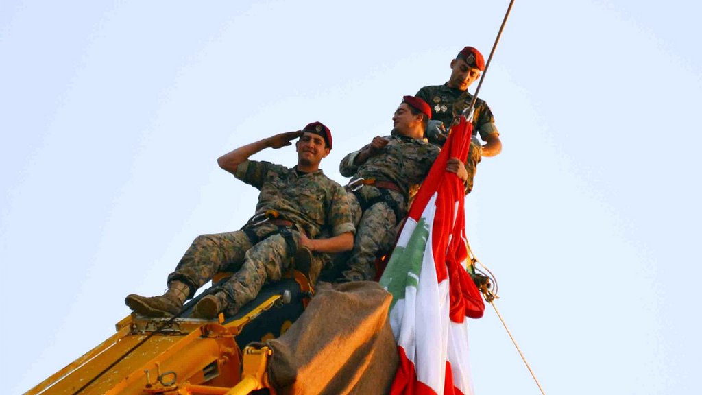 L'image montre trois soldats en uniforme, perchés en haut d'une grue. Ils sont en train de saluer tout en tenant un grand drapeau, probablement celui du Liban. Les soldats affichent un air déterminé et solennel, et l'ambiance semble festive, suggérant une célébration ou un événement important. Le ciel est clair, ce qui met en valeur la scène.