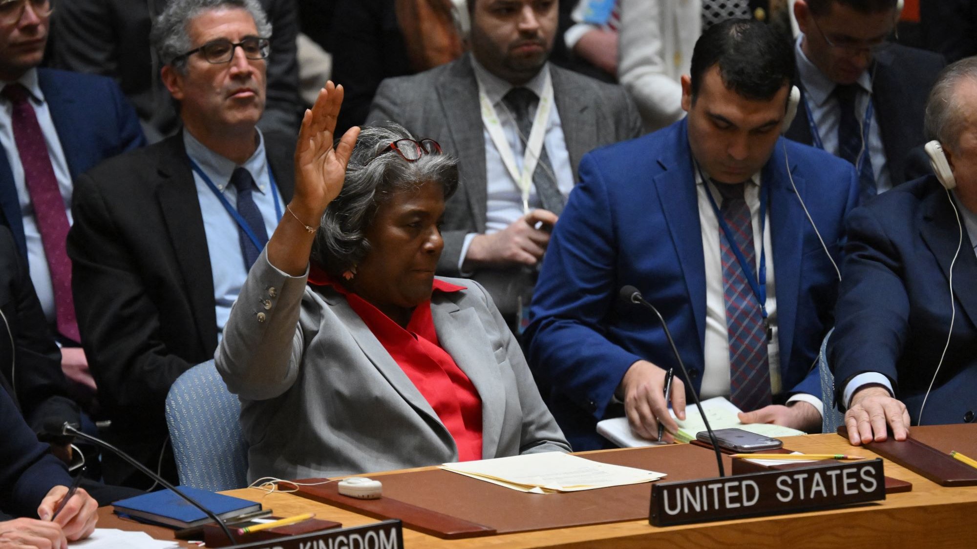 L'image montre une réunion au Conseil de sécurité des Nations Unies. Une femme, manifestement représentant les États-Unis, est assise à une table, levant la main pour prendre la parole ou voter. À côté d'elle, on aperçoit d'autres délégués de différents pays, tous attentifs. L'atmosphère semble sérieuse et concentrée, typique des discussions diplomatiques importantes.