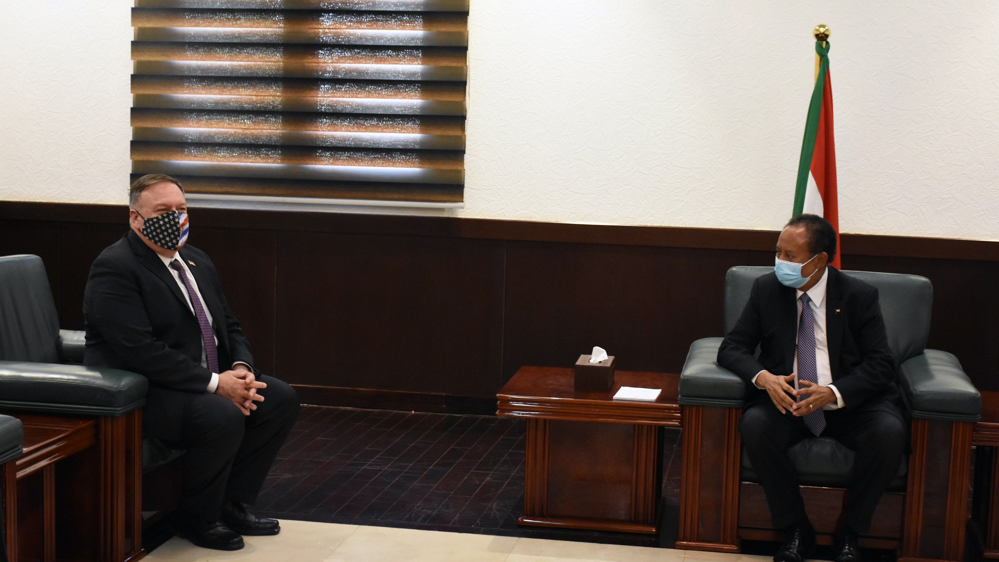 L'image montre deux hommes assis dans une pièce bien éclairée, avec des murs clairs et un sol en marbre. L'un des hommes porte un masque facial à motifs, tandis que l'autre est en costume avec une cravate. À l'arrière-plan, on peut voir un drapeau. Les deux hommes semblent engagés dans une conversation ou une réunion. Les meubles sont en bois sombre, et la pièce dégage une atmosphère formelle.