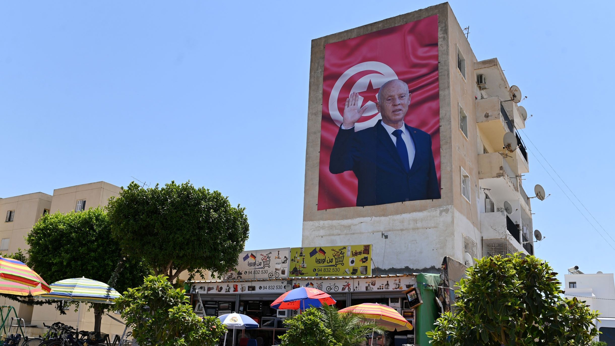L'image montre un bâtiment avec une grande fresque représentant un homme, probablement un dirigeant, qui fait un geste de salutation. En arrière-plan, on peut voir un drapeau tunisien. Au premier plan, il y a des parasols colorés et des arbres, ainsi qu'une scène de vie qui pourrait inclure des personnes assises ou se déplaçant. Le ciel est clair et ensoleillé, ce qui donne une atmosphère lumineuse à la scène.