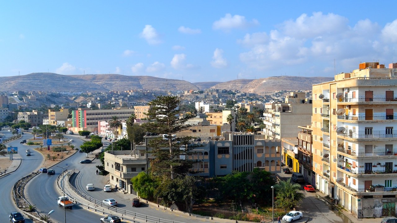 L'image montre un paysage urbain avec des bâtiments variés, y compris des immeubles résidentiels et commerciaux, ainsi qu'une route serpentine. Au loin, on peut voir des collines et un ciel clair avec quelques nuages. La scène dégage une ambiance paisible, avec des voitures circulant sur la route et des arbres ajoutant une touche de verdure à l'environnement.