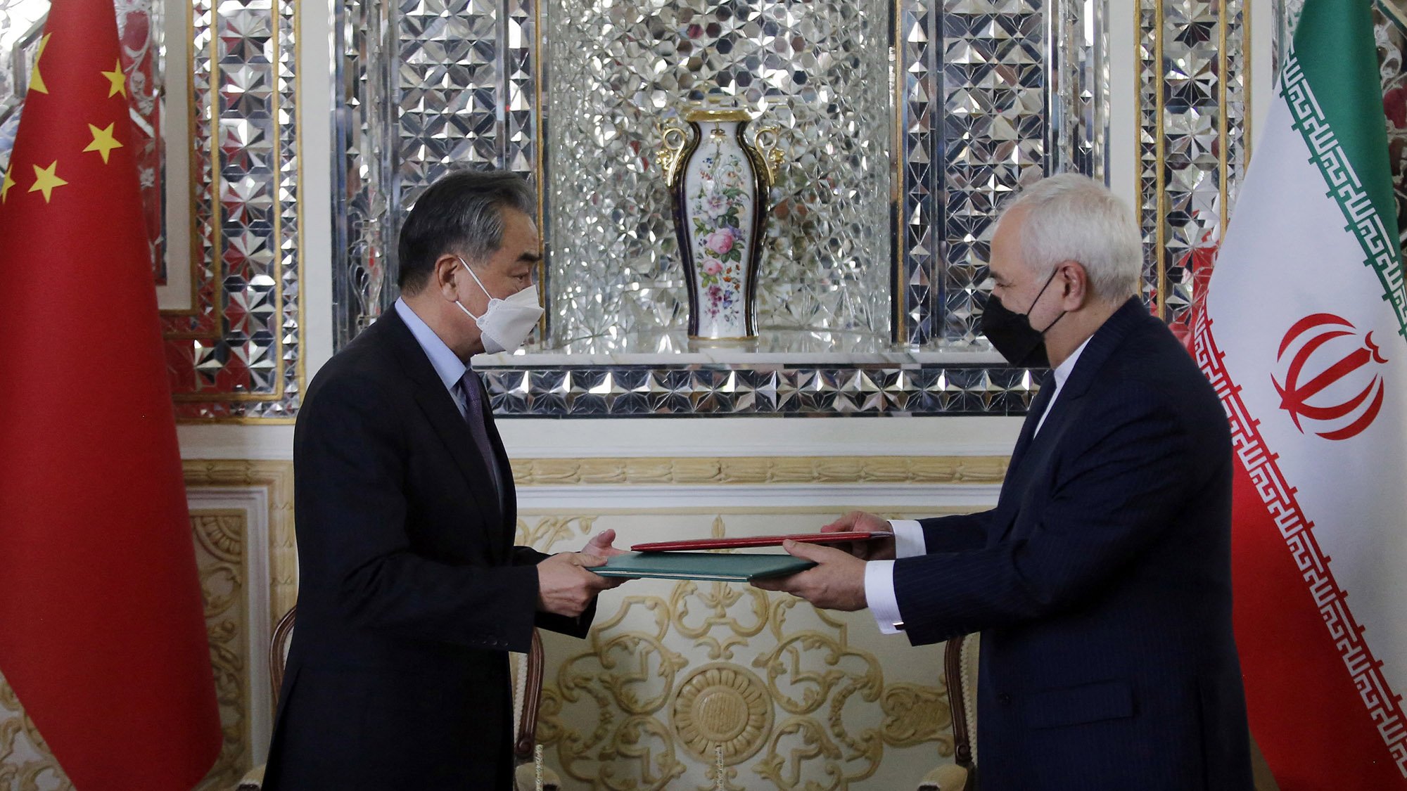 L'image montre deux hommes, apparemment des représentants de la Chine et de l'Iran, se tenant en face l'un de l'autre. Ils échangent des documents au cours d'une cérémonie formelle. On peut voir les drapeaux de la Chine et de l'Iran en arrière-plan. L'environnement est richement décoré, avec des motifs élaborés et un vase floral visible derrière eux. Les deux hommes portent des masques, ce qui pourrait suggérer que la photo a été prise pendant la pandémie.