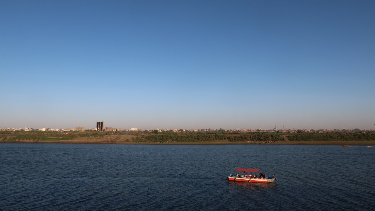L'image montre une scène paisible d'un cours d'eau avec un ciel dégagé et sans nuages. Au centre, on peut voir un petit bateau naviguant sur les eaux calmes. À l'arrière-plan, on distingue une ligne d'horizon urbaine, avec des bâtiments et des arbres le long de la rive. L'ambiance générale est tranquille et ensoleillée, suggérant une belle journée.