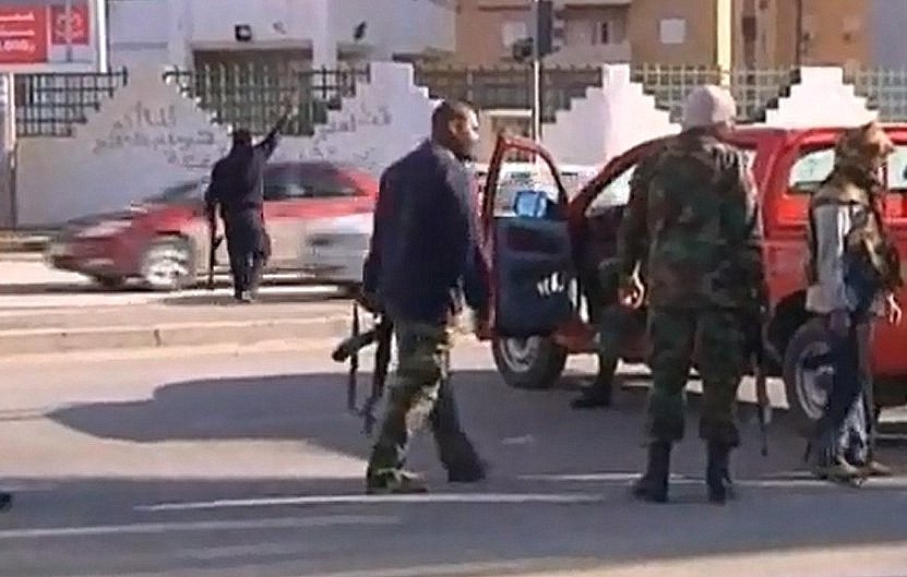 L'image montre une scène de chaos impliquant des personnes en uniforme et armées. Quelques individus se déplacent près de véhicules, tandis qu'un homme semble gesticuler au loin. L'environnement urbain est visible, avec des bâtiments en arrière-plan et une circulation routière. L'atmosphère semble tendue, suggérant un contexte de conflit ou d'incident de sécurité.