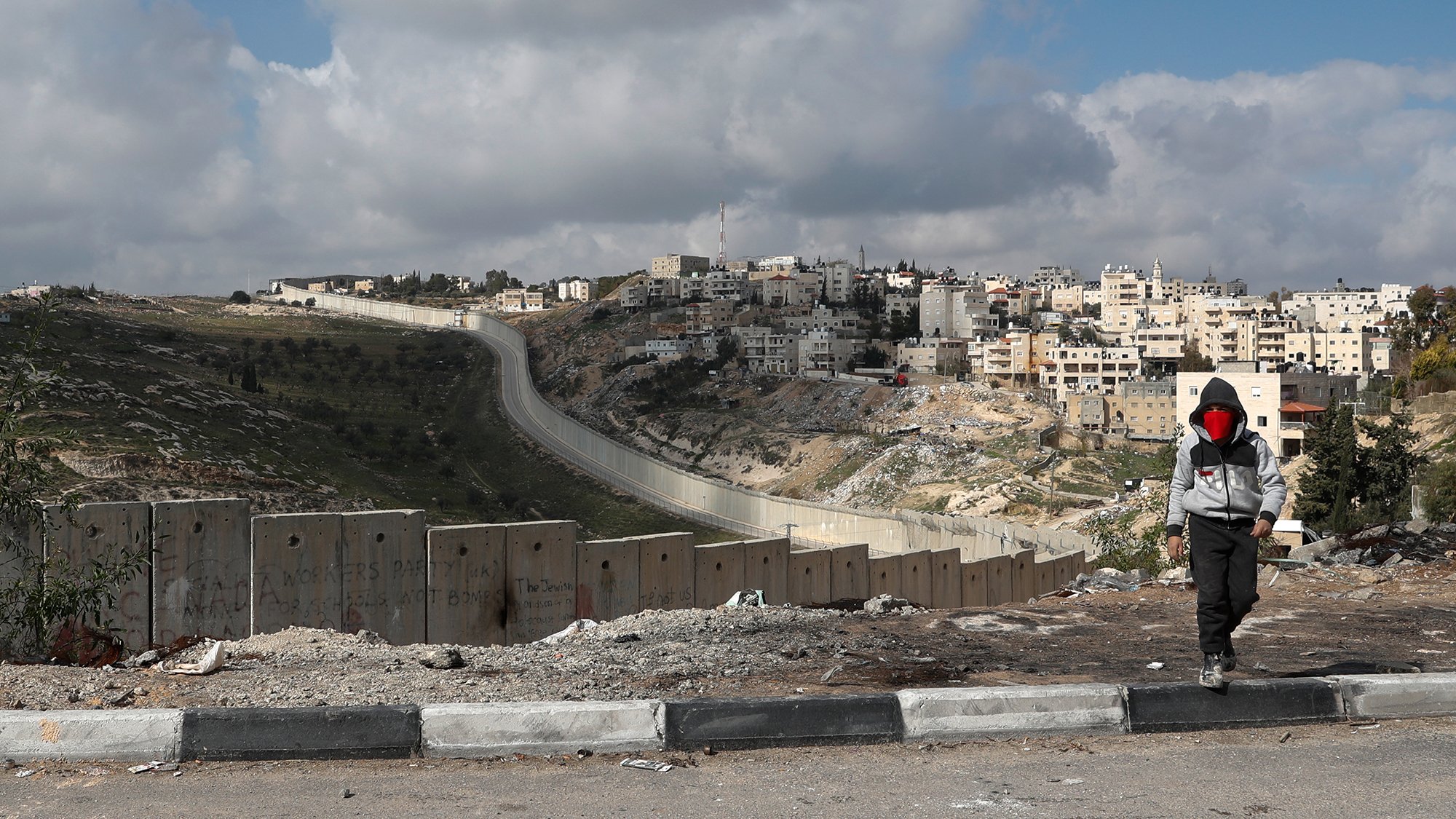 L'image montre un enfant marchant le long d'une route, avec des blocs de béton formant une barrière sur le côté. En arrière-plan, on peut voir une colline surmontée de bâtiments, indiquant une zone urbaine. Le ciel est partiellement nuageux, et le paysage semble aride et rocailleux. L'enfant porte une capuche et un masque, ajoutant une dimension de mystère à la scène. La barrière suggère des thèmes de division et de conflit.