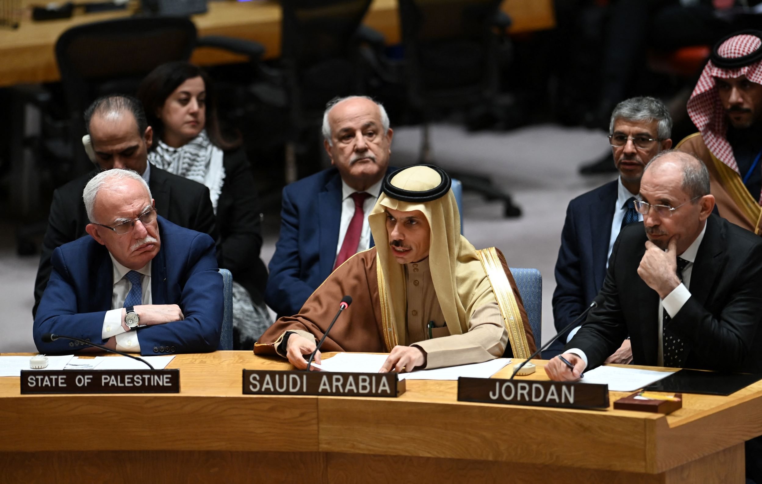 L'image montre une réunion de l'Assemblée générale ou du Conseil de sécurité de l'ONU. Plusieurs représentants de différents pays sont assis autour d'une table. À gauche, on voit un homme portant des lunettes avec un badge indiquant "État de Palestine". Au centre, un homme en tenue traditionnelle saoudienne prend la parole, tandis qu'un autre à droite, en costume noir, représente la Jordanie. En arrière-plan, d'autres participants écoutent attentivement. L'atmosphère semble sérieuse, suggérant une discussion importante.