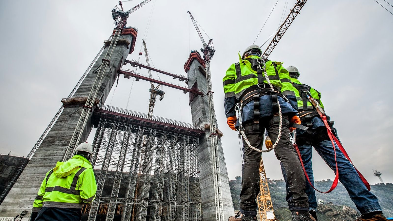 L'image montre une scène de construction avec plusieurs travailleurs vigilants observant une structure imposante en cours d'édification. Les ouvriers portent des vestes de sécurité à rayures et des casques, et sont accrochés avec des harnais de sécurité. En arrière-plan, on peut voir des grues et des éléments de construction en cours d'assemblage, tandis que le ciel est nuageux, suggérant une ambiance de travail intense sur le chantier.