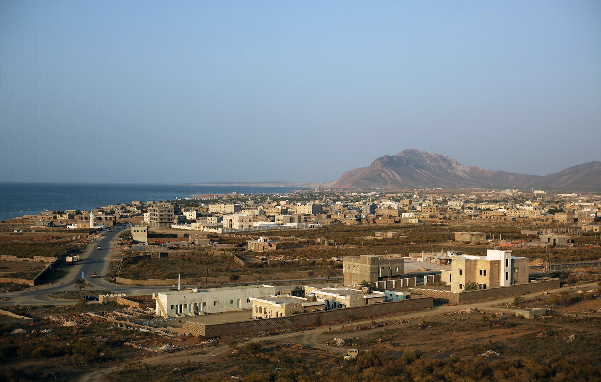 L'image montre un paysage côtier avec une vue sur une petite ville située près de la mer. On peut apercevoir des bâtiments de différentes tailles, certains modernes et d'autres plus traditionnels, dispersés sur une étendue de terrain. À l'arrière-plan, on distingue une montagne dont les contours se dessinent sous un ciel clair. La route serpente à travers la ville, menant à la mer. L'ensemble dégage une atmosphère paisible et semi-aride, typique des régions côtières.