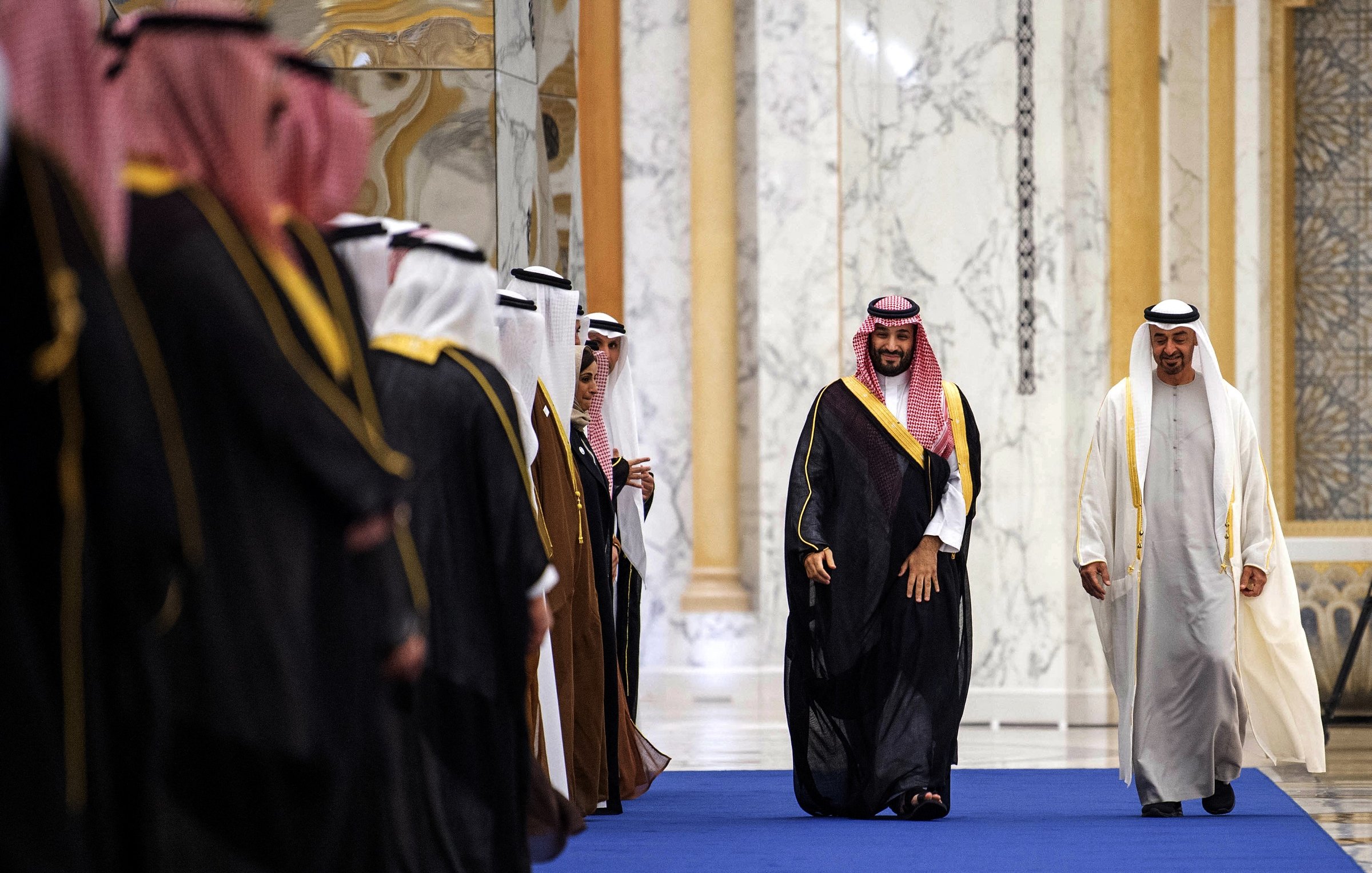 L'image montre un groupe d'hommes vêtus de vêtements traditionnels, probablement en Arabie Saoudite ou dans un pays du Golfe. Leurs tenues incluent des robes longues et des châles sur la tête. Au centre, un homme en particulier se distingue, souriant et avançant sur un tapis bleu. Il semble être en train d'interagir avec les autres hommes qui se tiennent de part et d'autre, dans un décor élégant avec des murs en marbre. L'ambiance semble formelle et festive.