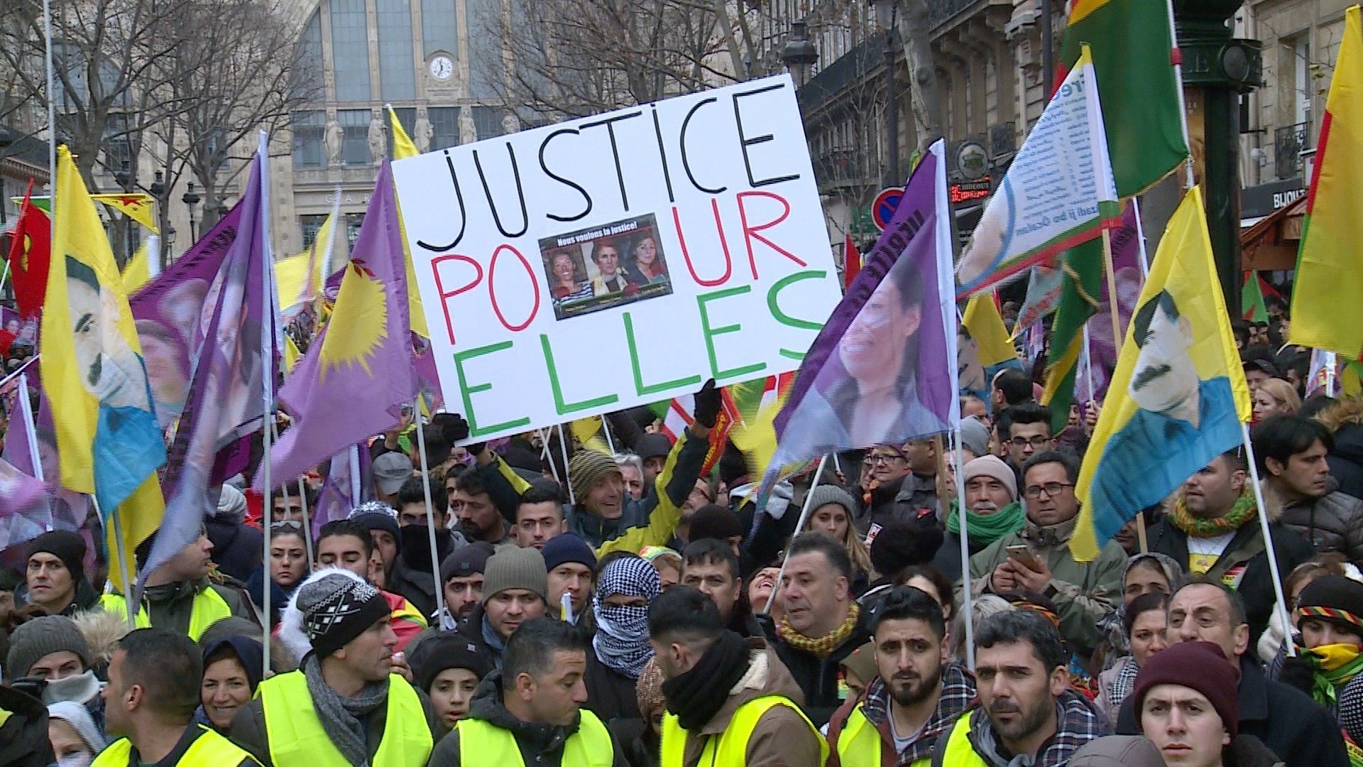 L'image montre une manifestation avec de nombreuses personnes rassemblées. Les manifestants portent des drapeaux colorés et des panneaux. L'un des panneaux affiche le message "JUSTICE POUR ELLES", suggérant un appel à la justice, probablement pour des femmes. Les visages des manifestants sont déterminés, et l'atmosphère semble impliquée et engagée.
