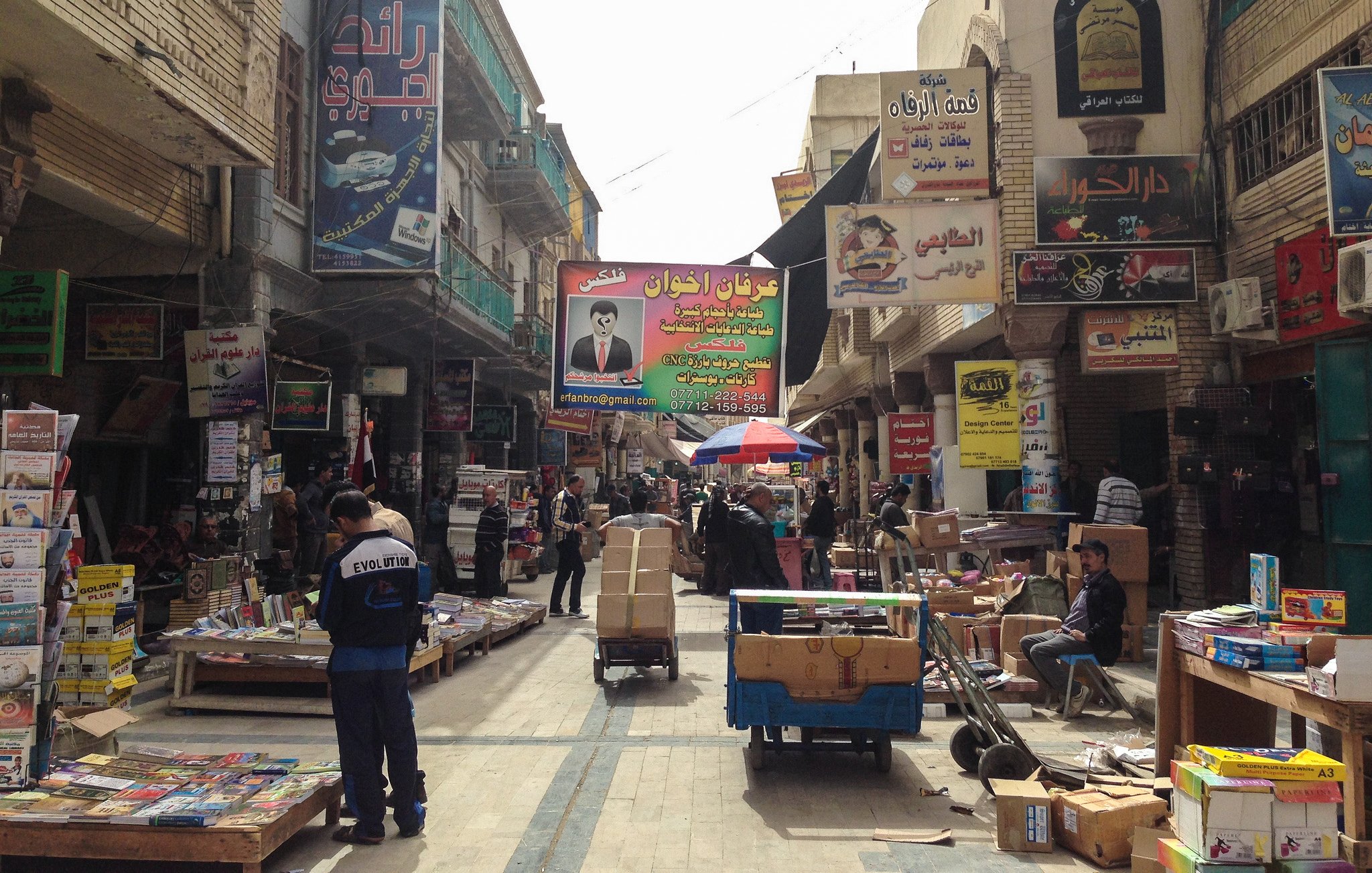 L'image montre une rue animée, typique d'un marché urbain dans une ville. On peut voir des étals et des magasins de part et d'autre, présentant divers produits, avec des affiches et des panneaux publicitaires colorés accrochés aux murs. Certaines personnes sont visibles, allant et venant, tandis que d'autres semblent être en train de travailler, probablement en déchargeant des marchandises. Le sol est souvent encombré de caisses et d'autres objets, créant une ambiance vivante et dynamique. Des parasols sont également présents, offrant un peu d'ombre dans cette rue fréquentée.