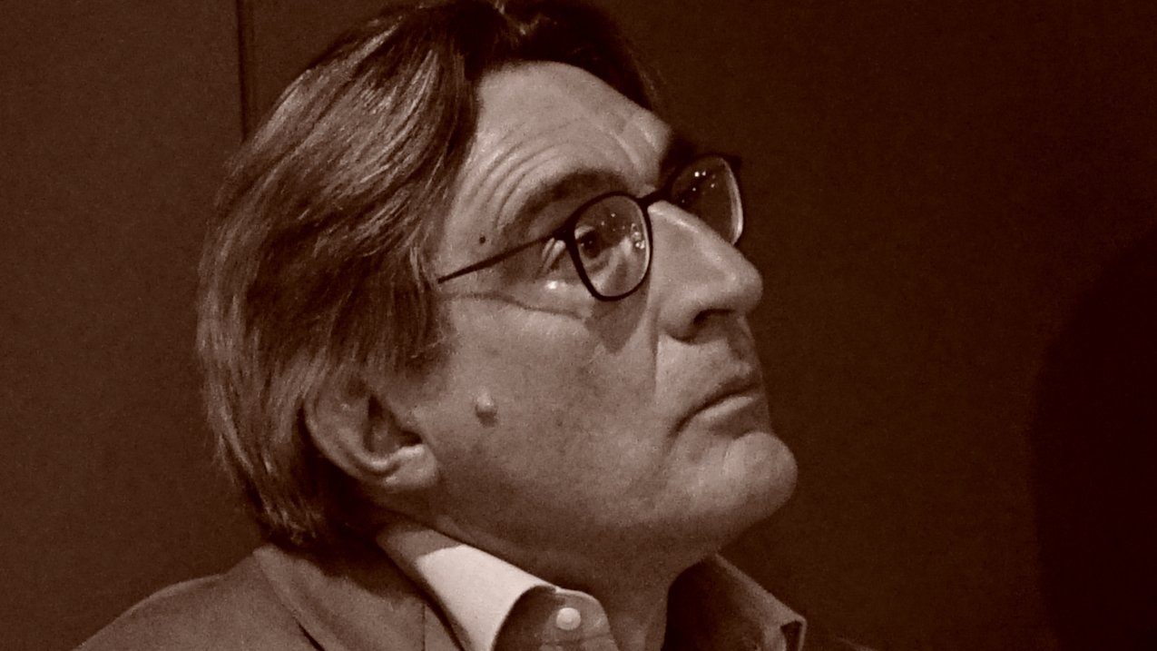 L'image montre un homme aux cheveux grisonnants, portant des lunettes et un costume clair. Il semble pensif, regardant quelque chose hors champ. L'éclairage et le ton de l'image lui donnent un aspect rétro, suggérant une ambiance contemplative.