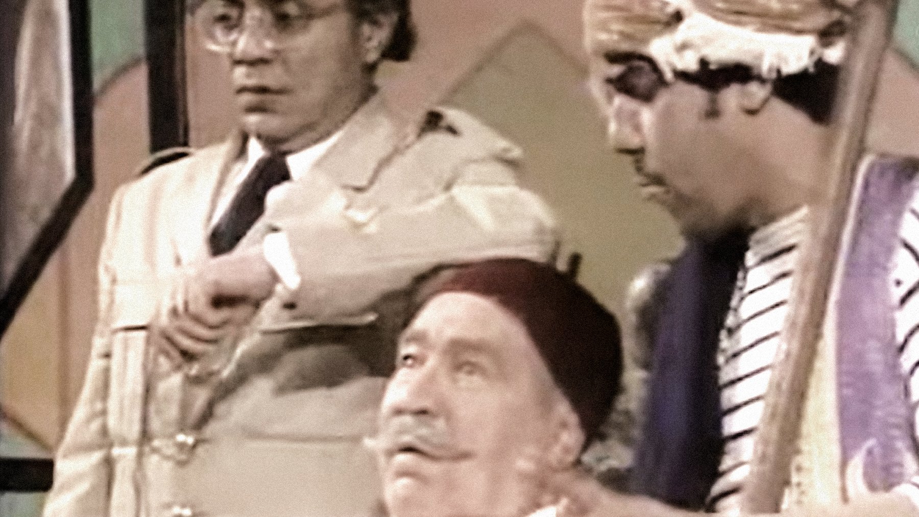 L'image montre trois personnages dans un cadre qui semble être une scène de théâtre ou une comédie. L'un des personnages est assis, portant une blouse blanche et un fez, affichant une expression détendue. À sa gauche se trouve un personnage en tenue militaire, regardant sa montre avec une expression préoccupée. À droite, un troisième personnage, également en fez, joue d'un instrument de musique tout en gesticulant, ajoutant une ambiance dynamique à la scène. L'ensemble évoque un moment comique ou dramatique.