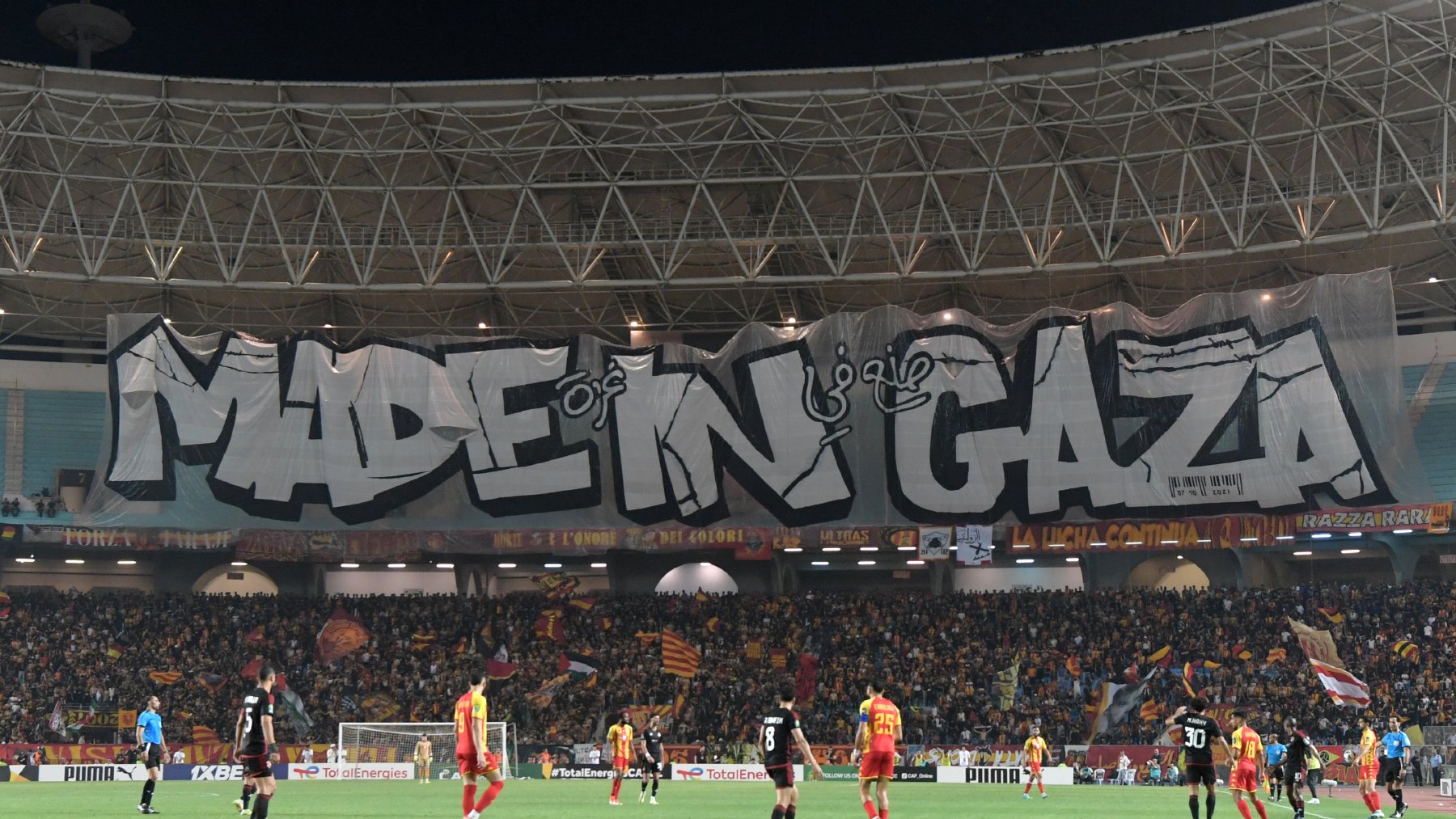 L'image montre un stade de football avec une grande banderole affichée. La banderole est ornée du texte "MADE IN GAZA" écrit en lettres stylisées et imposantes. Le stade est rempli de supporters, et l'atmosphère semble animée. On peut voir des joueurs sur le terrain, ce qui indique qu'un match est en cours. L'ambiance est festive et engagée.