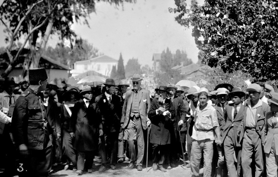 L'image montre un groupe de personnes rassemblées en extérieur, avec une scène animée. Au centre, une figure éminente se déplace, soutenue par une canne. De nombreuses personnes autour d'elle portent des chapeaux et tiennent des parasols, probablement pour se protéger du soleil. Des hommes en costume se distinguent parmi la foule, et l'environnement semble être urbain, avec des bâtiments en arrière-plan. La scène dégage une atmosphère d'importance ou de célébration.