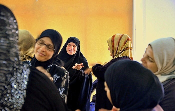 L'image montre un groupe de femmes en discussion ou en interaction. Elles portent des vêtements traditionnels, y compris des hijabs et des abayas. Certaines femmes sourient et semblent engagées dans une conversation joyeuse. L'éclairage dans la pièce donne une ambiance chaleureuse, avec un fond qui semble jaune. L'atmosphère générale est conviviale et animée.