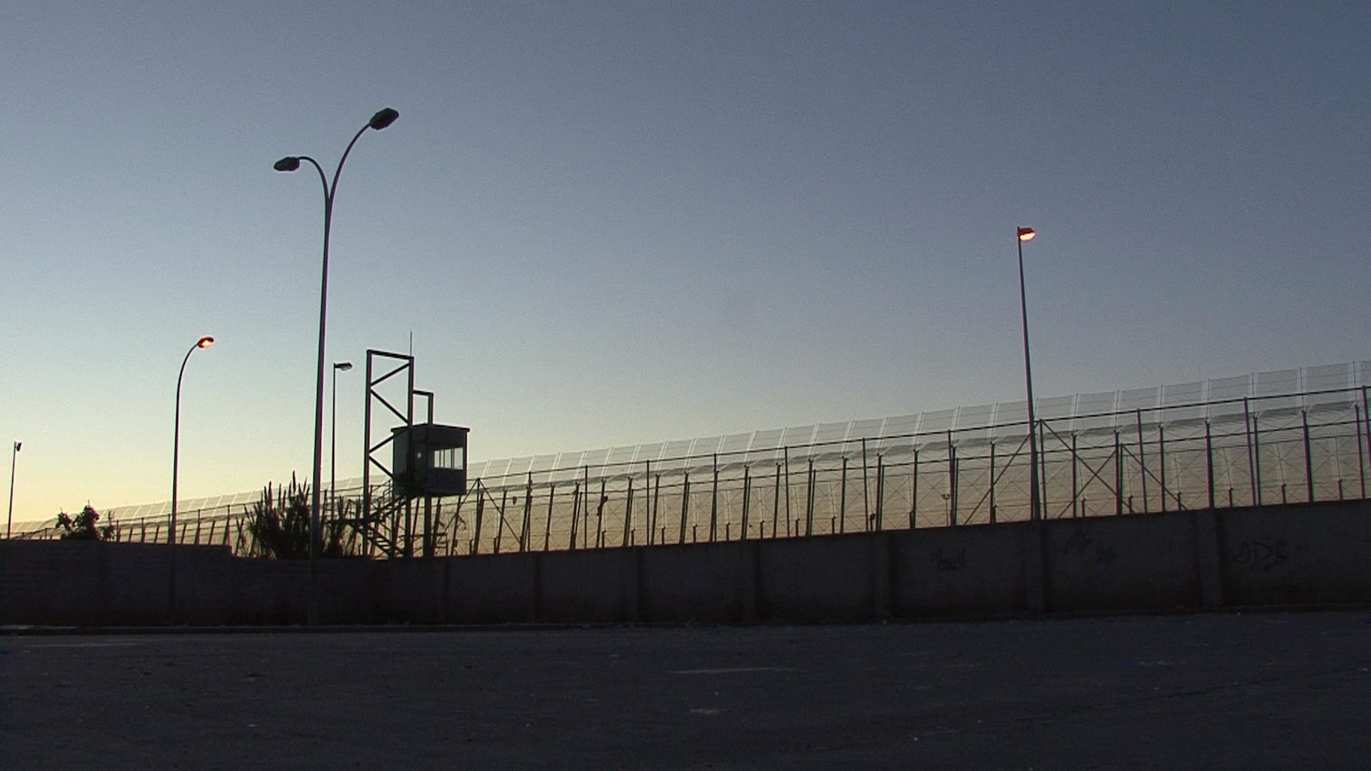 L'image montre une clôture de sécurité, probablement d'une prison ou d'une zone contrôlée. On aperçoit une tour d'observation au fond, illuminée par des lampadaires. La scène se déroule au crépuscule, avec un ciel qui passe des teintes orange et bleues, créant une atmosphère à la fois calme et tendue. Le mur en béton à la base de la clôture souligne le caractère restrictif de l'endroit.