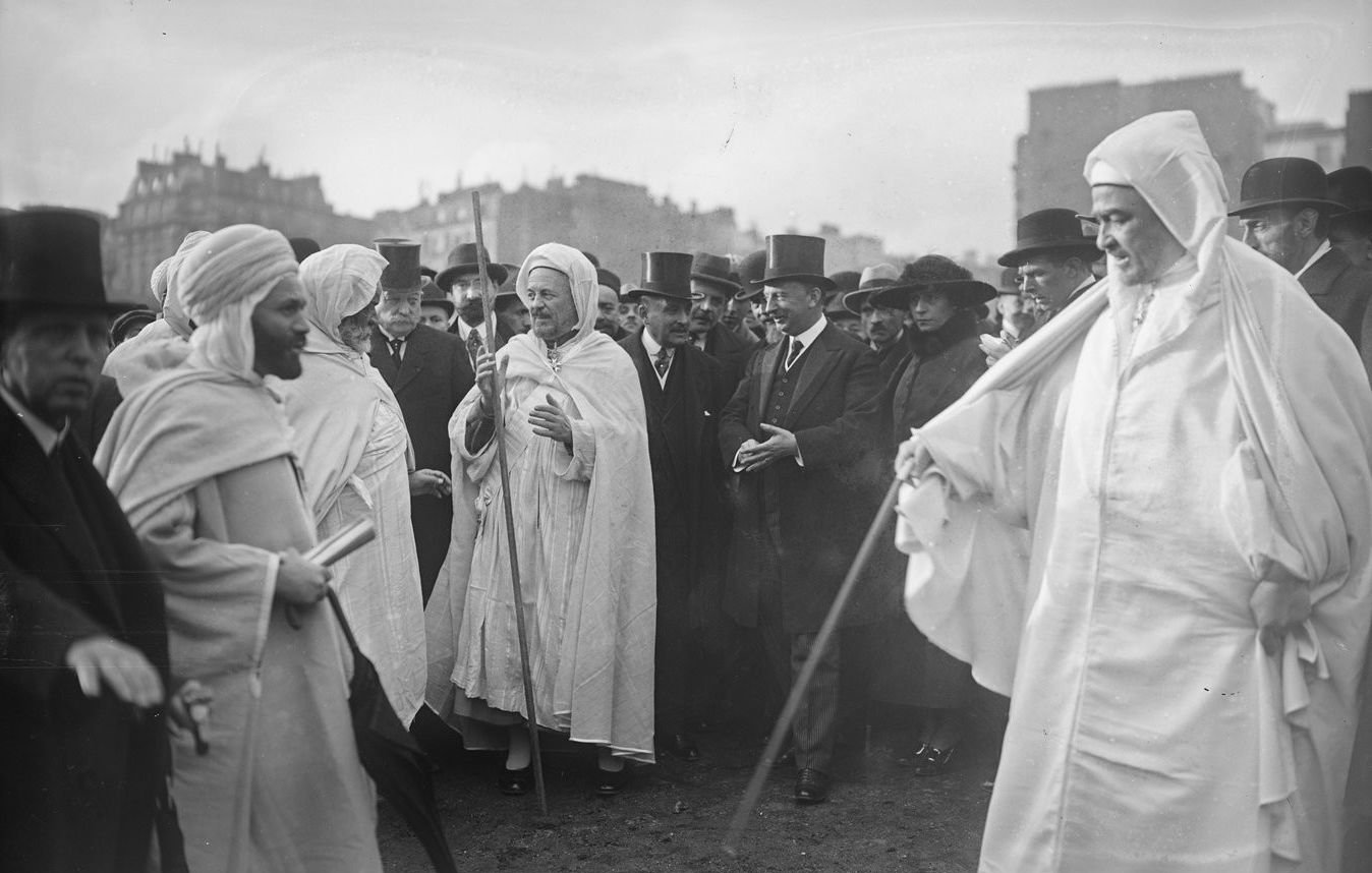 L'image montre un groupe de personnes dans un environnement urbain. Au centre, plusieurs individus portent des vêtements traditionnels, tels que des robes blanches et des turbans. D'autres, habillés de manière formelle avec des chapeaux haut de forme et des costumes, se tiennent autour d'eux, suggérant une rencontre entre différentes cultures. L'arrière-plan présente des bâtiments typiques de l'époque. L'atmosphère semble être celle d'un événement officiel ou d'une cérémonie.