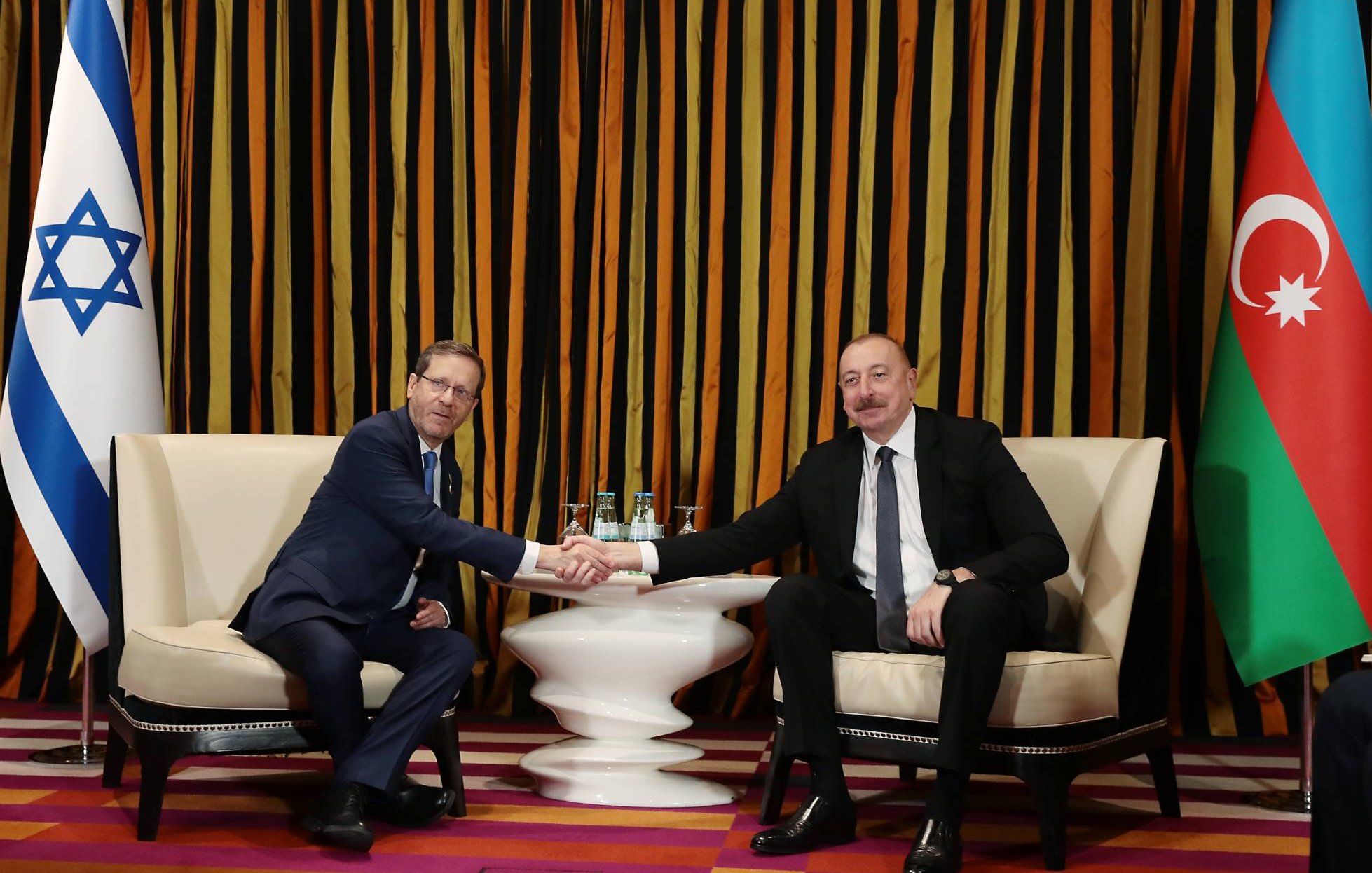 Deux hommes, en costume, se serrent la main dans un cadre élégant avec des drapeaux.